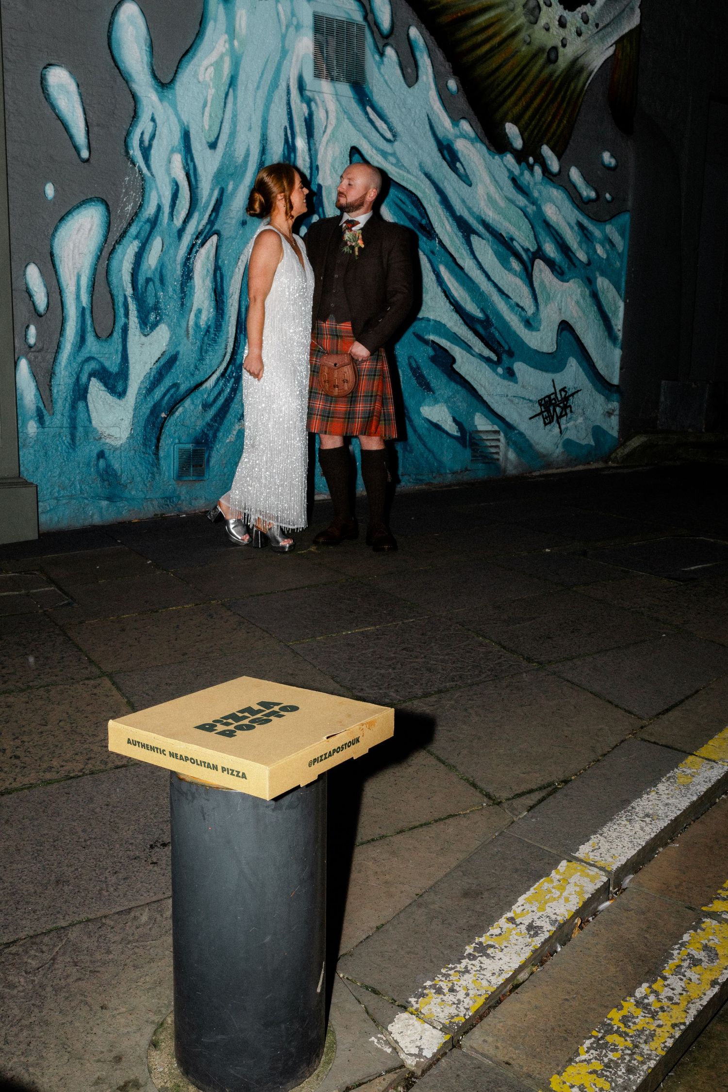 Scottish wedding couple kissing by colorful graffiti wall at night with pizza box in foreground.