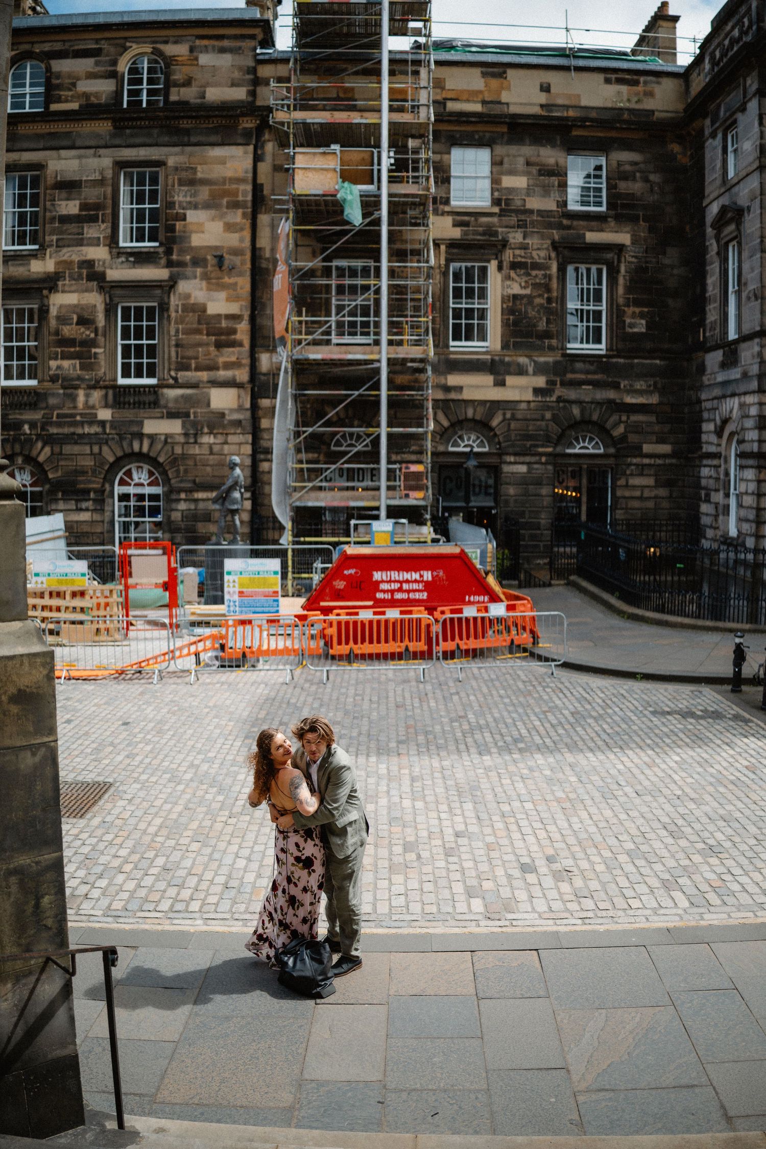 Bride and groom embrace on cobblestone square outside historic Edinburgh stone building.