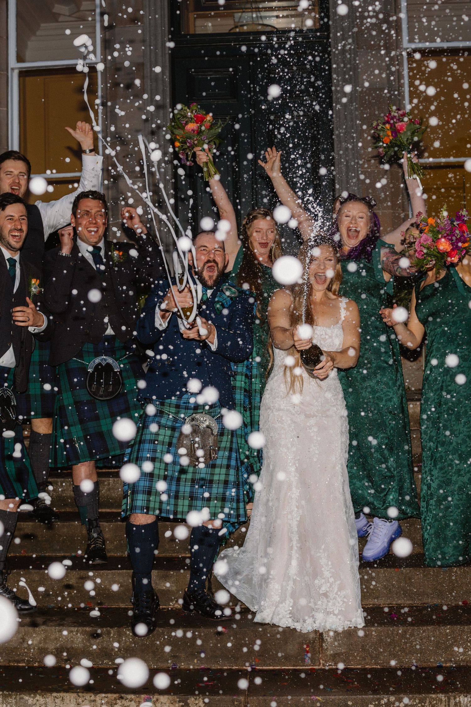 Bride and groom celebrate with wedding party on steps as bubbles fill the night air in joyful exit.