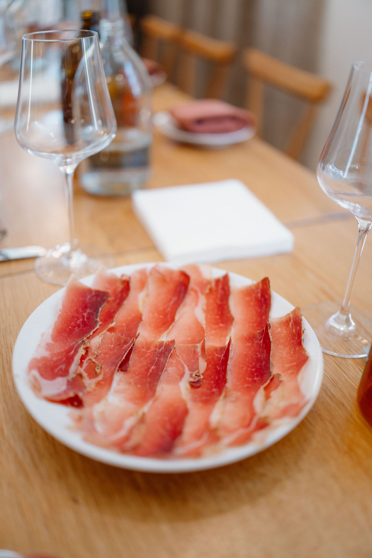Thinly sliced prosciutto arranged on a white plate beside empty wine glasses on a wooden table.