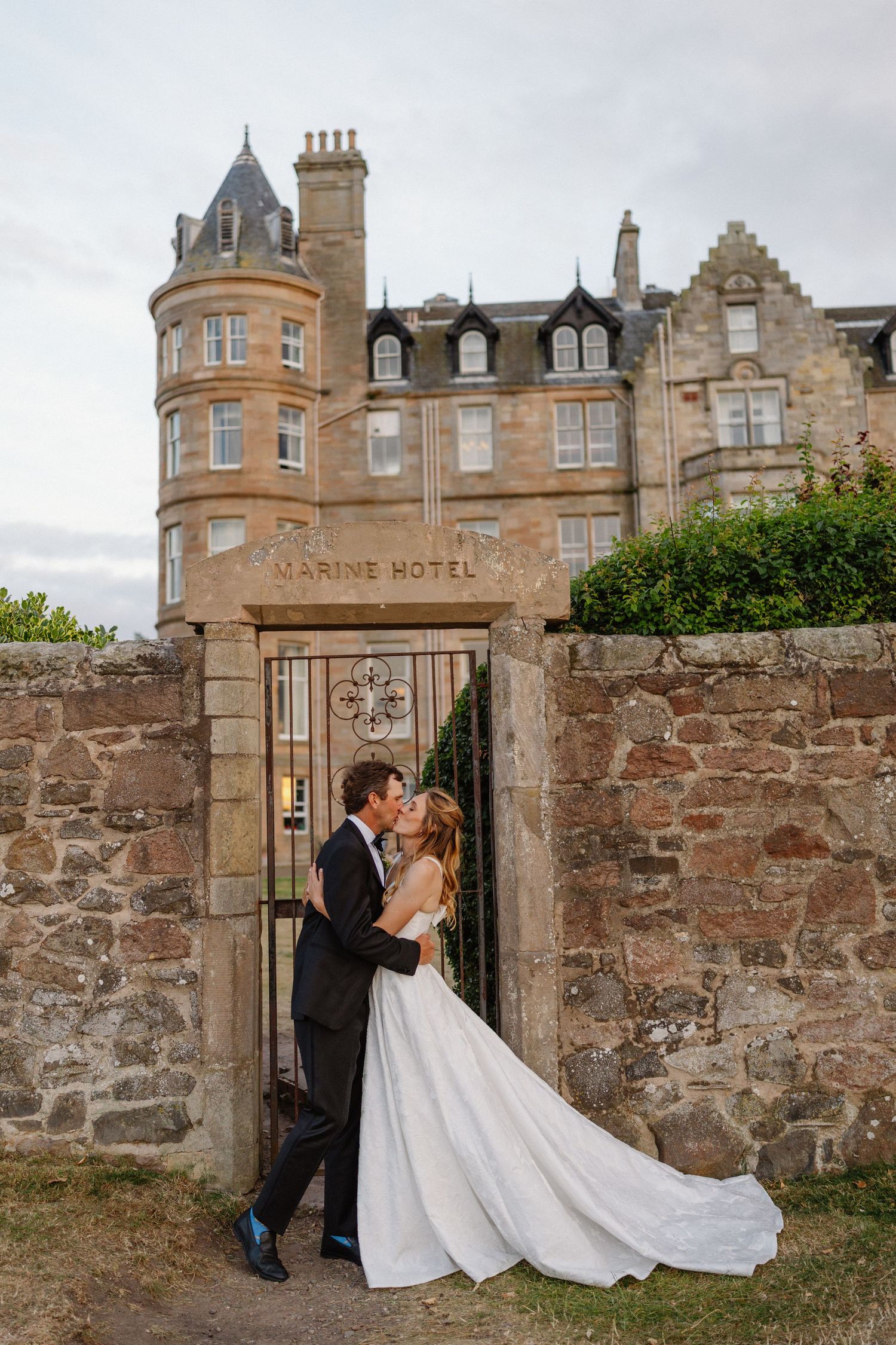Wedding couple kissing at ornate gate of Marine Hotel, bride in flowing white gown with long train.