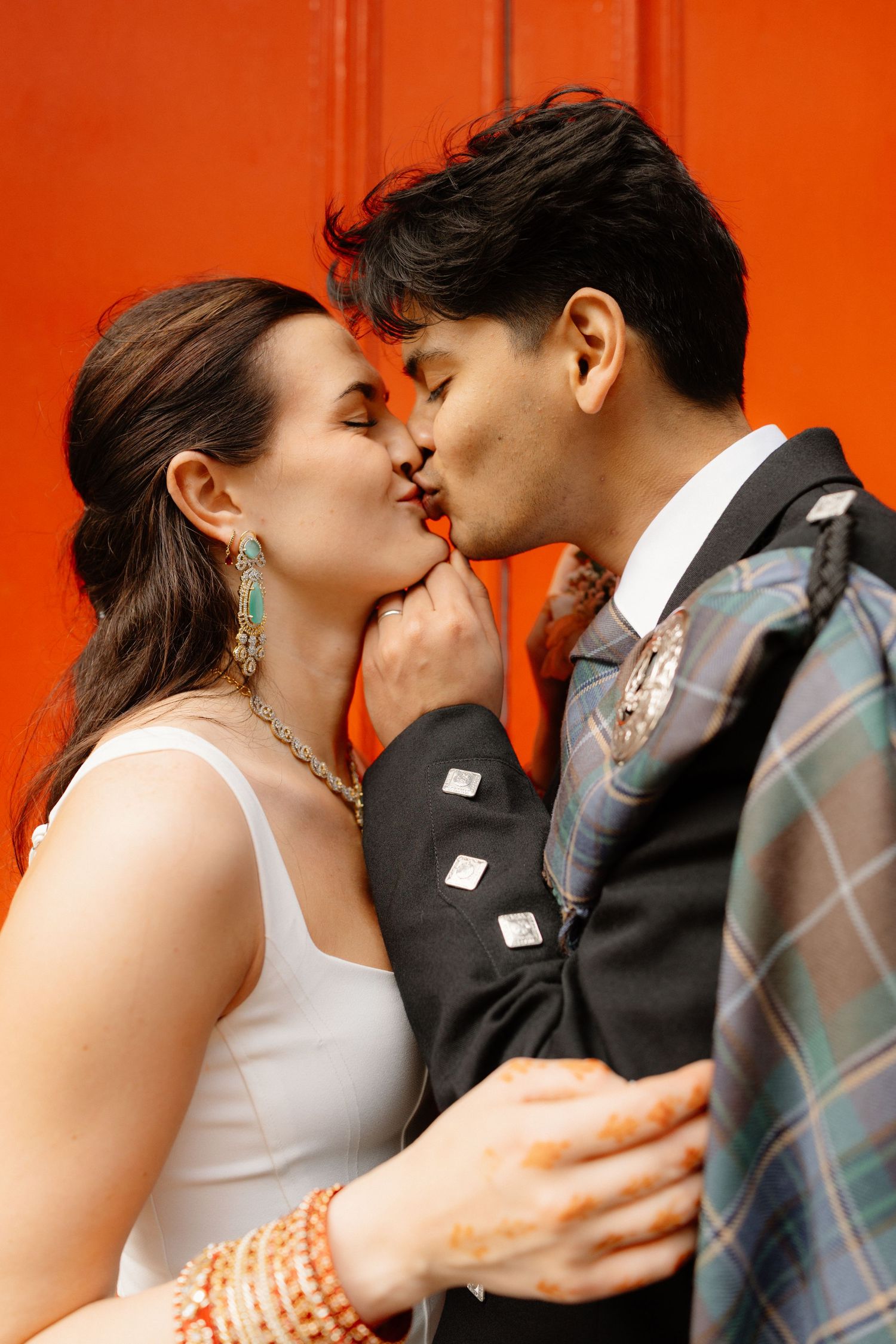 Bride and groom sharing a kiss against a vibrant red door, groom wearing a traditional Scottish tartan kilt.