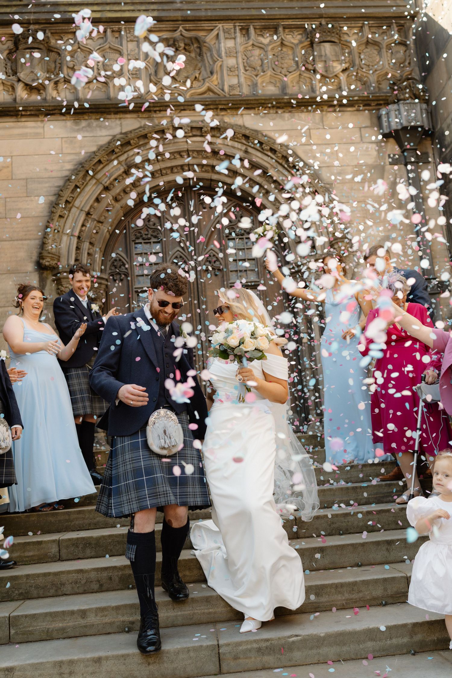 Bride and groom exit church in confetti shower, groom wearing traditional Scottish kilt.