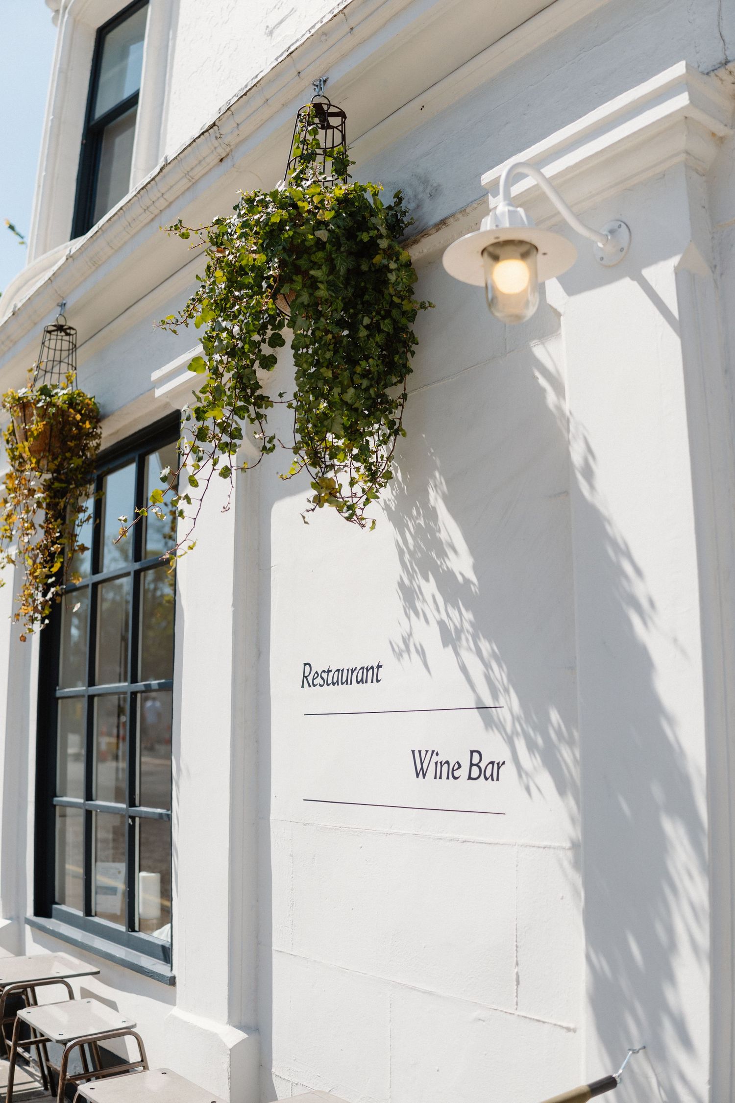 White exterior of a wine bar and restaurant with hanging plants and large windows.