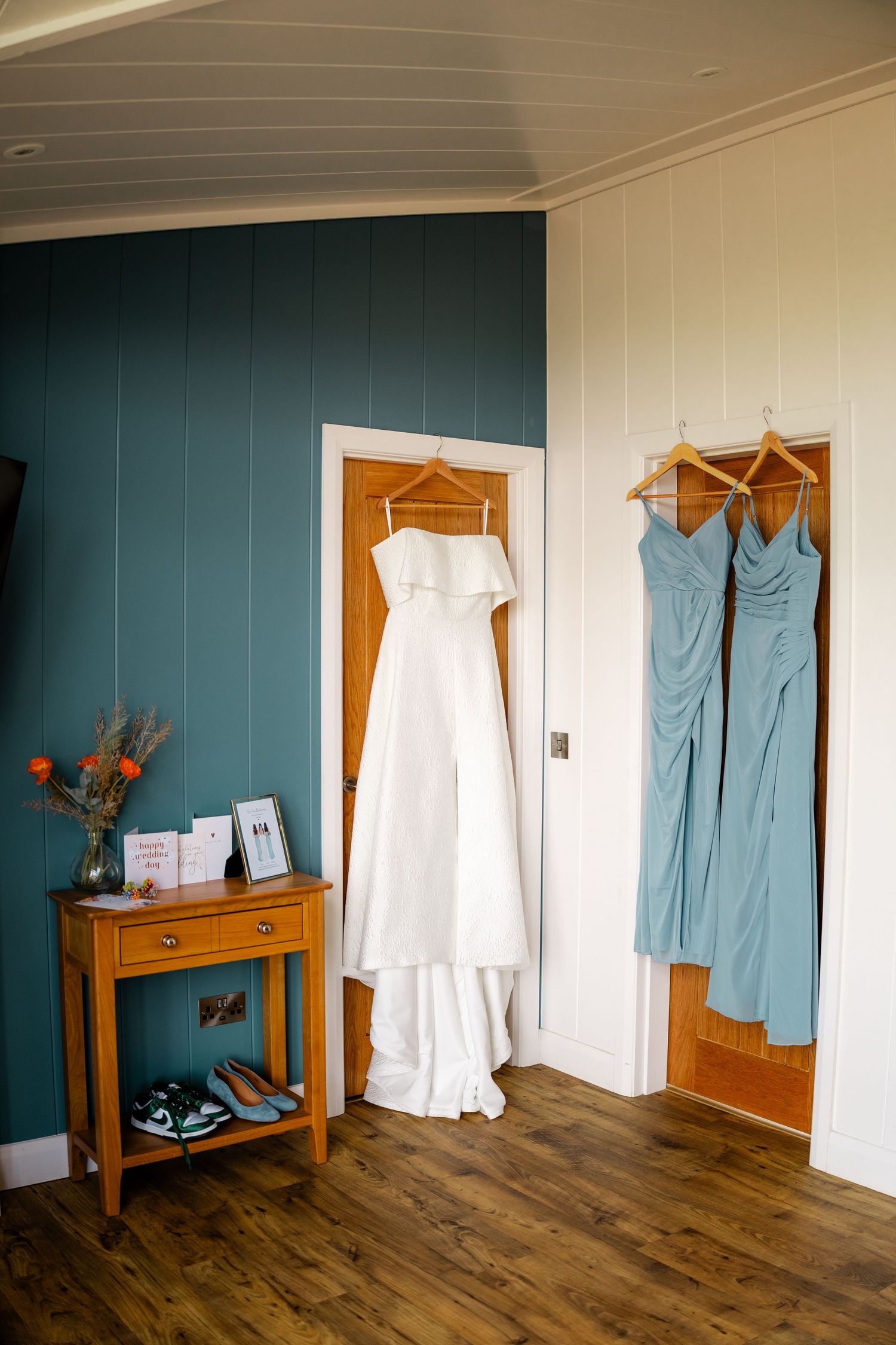 Wedding attire display showing white dress and blue bridesmaid gowns hanging in a room with wooden floors.
