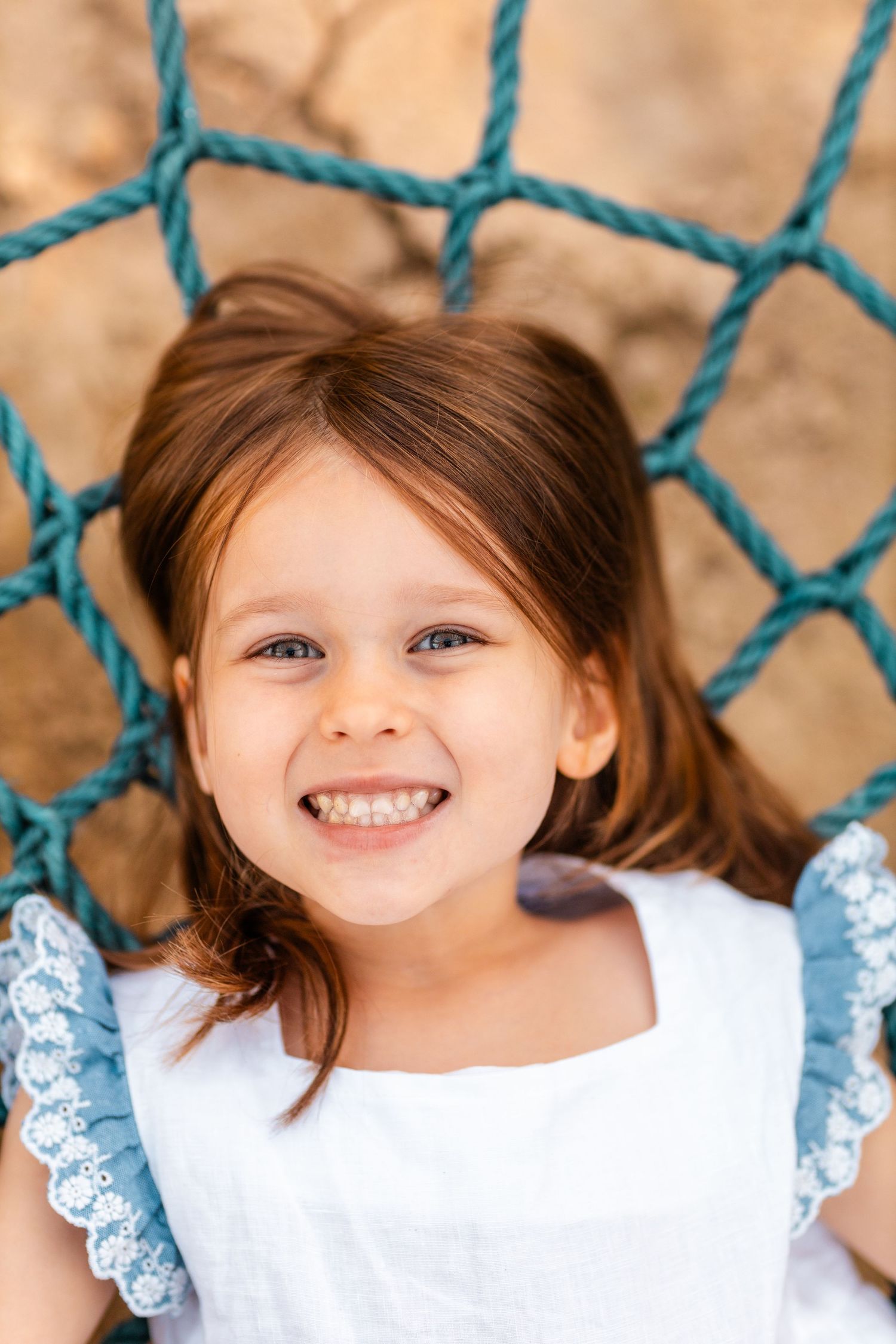 A young child in a white lace dress smiles while posing against blue ropes.