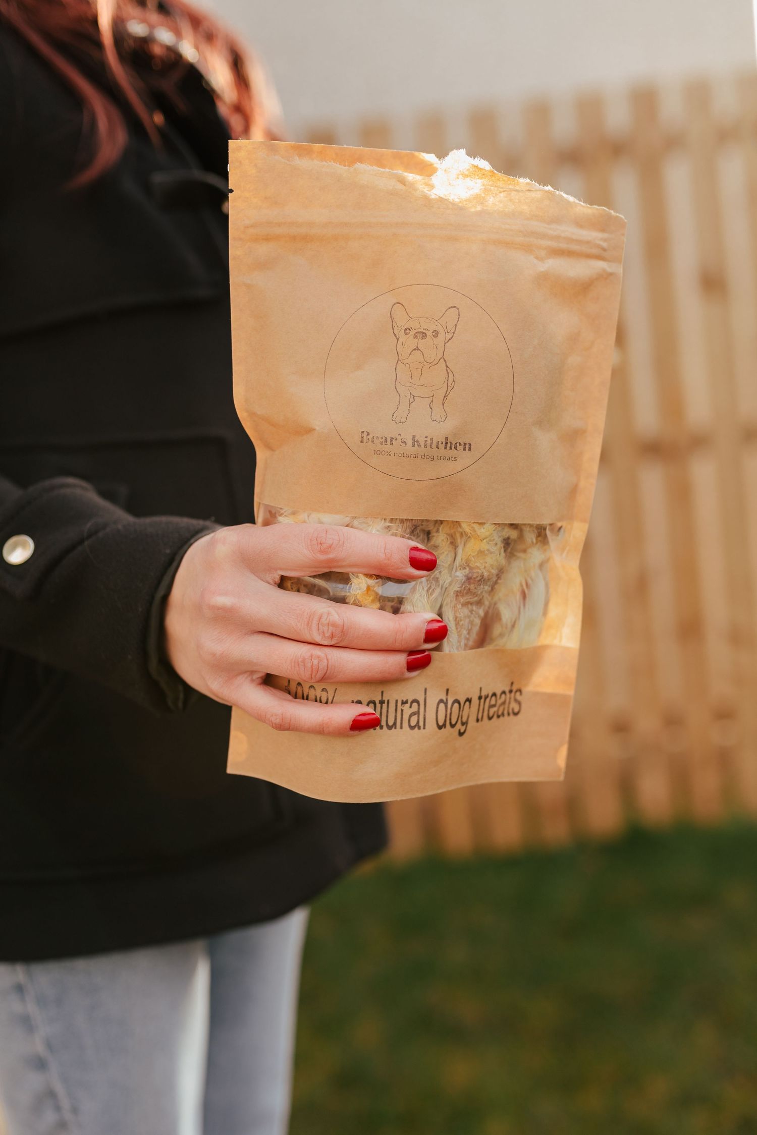 Hands holding natural dog treat paper bag outdoors with fence in background.