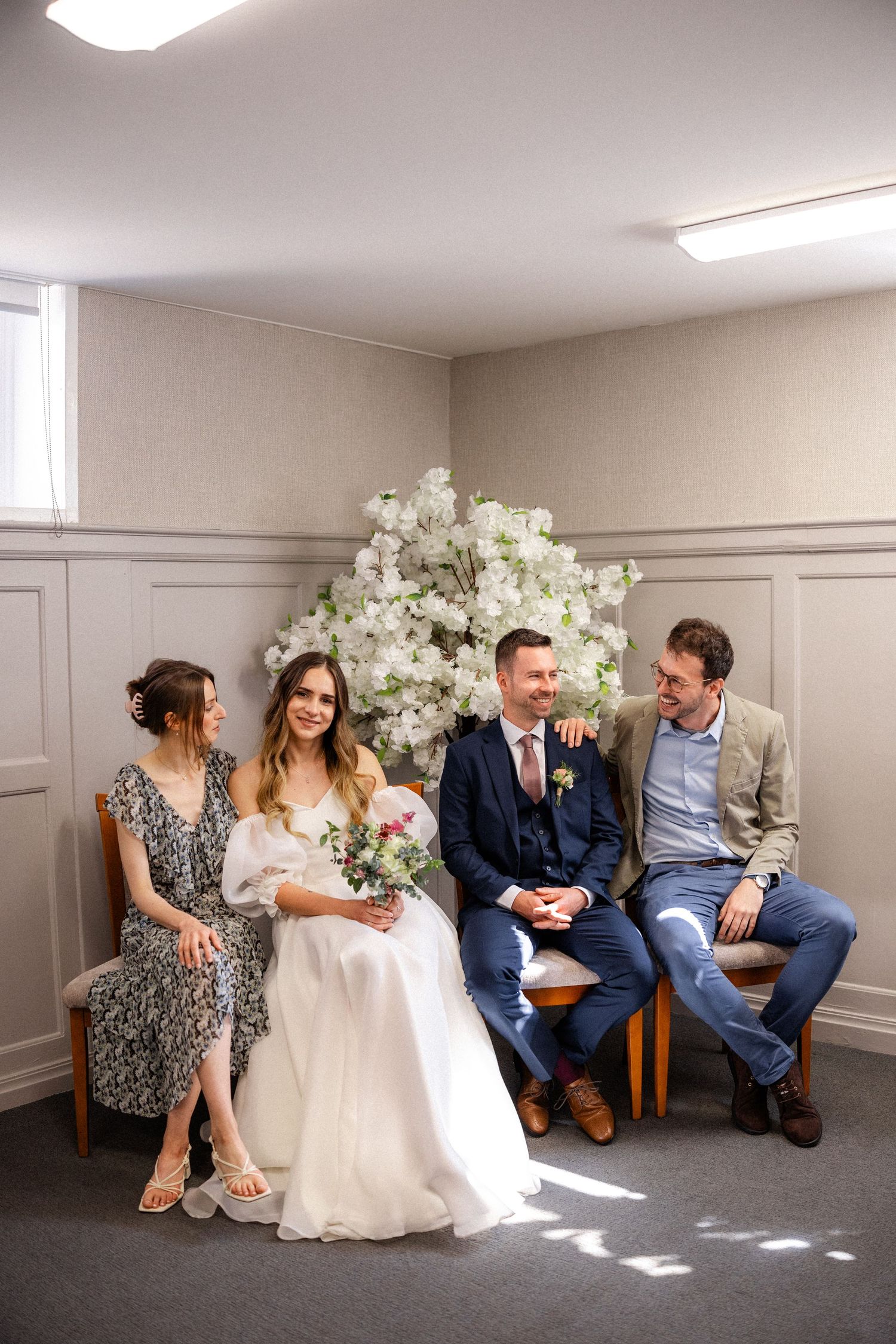 Wedding party relaxes on chairs in a bright, minimalist room with white floral decorations.