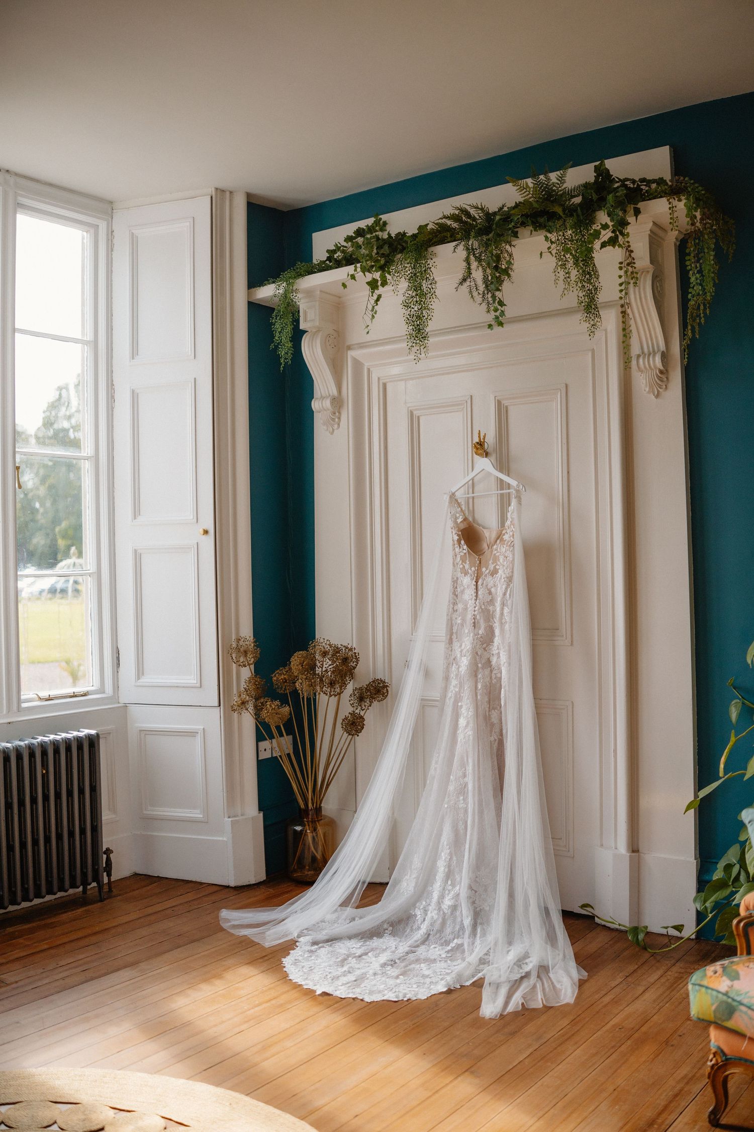 Wedding dress displayed in a bright room with white paneled walls, hardwood floors, and hanging greenery decorations.