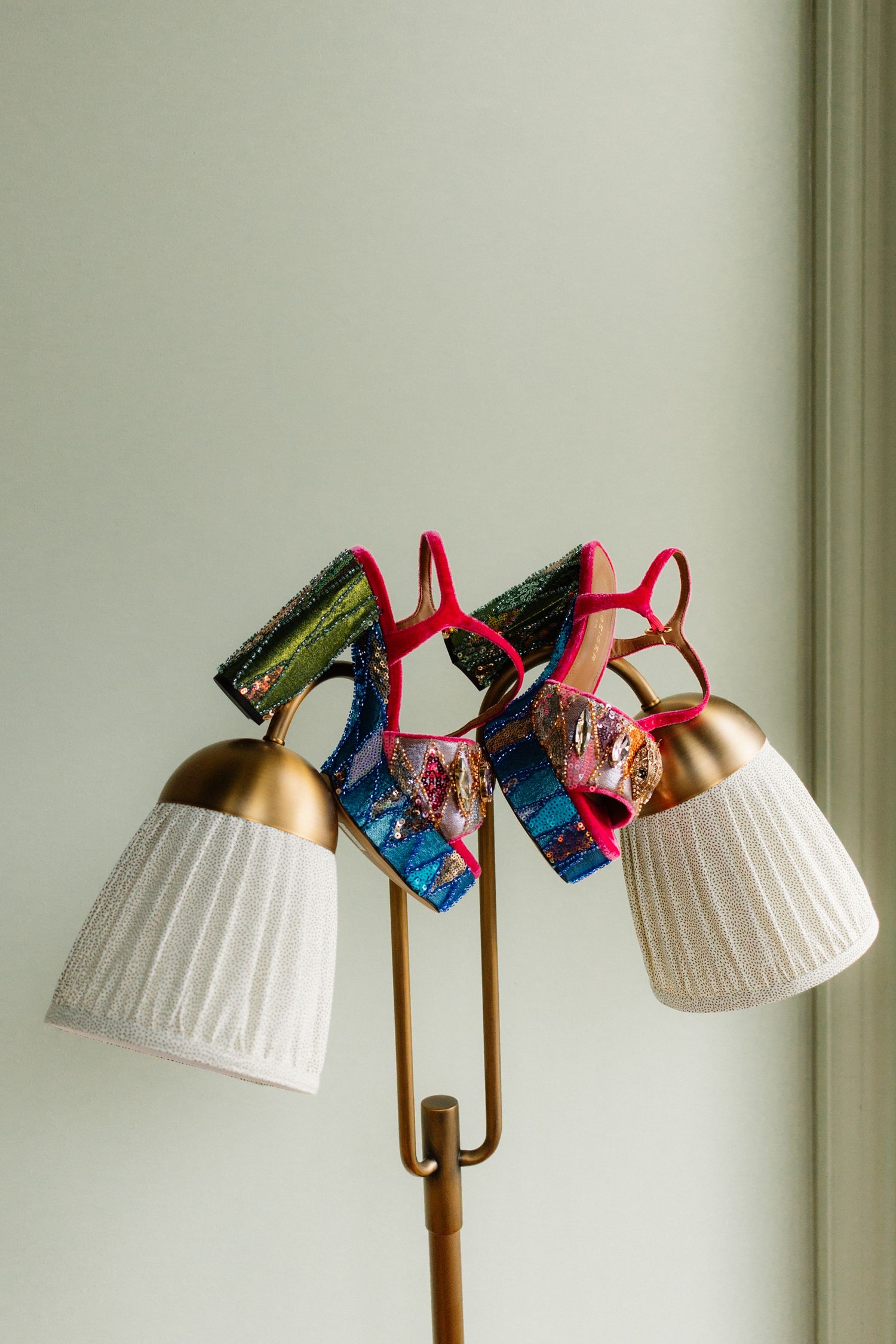 Colorful embellished high-heel sandals displayed on a brass floor lamp with white pleated shades.