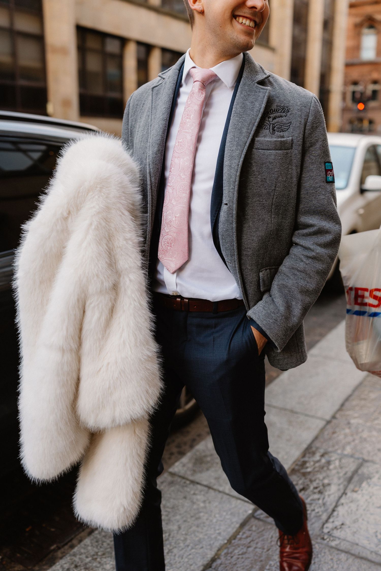 Smiling man in gray blazer, pink tie, and navy pants carries a white fur coat on a city street.