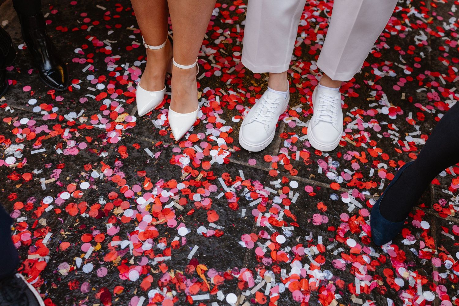 Two people in white shoes standing on colorful confetti-covered ground at a wedding celebration.