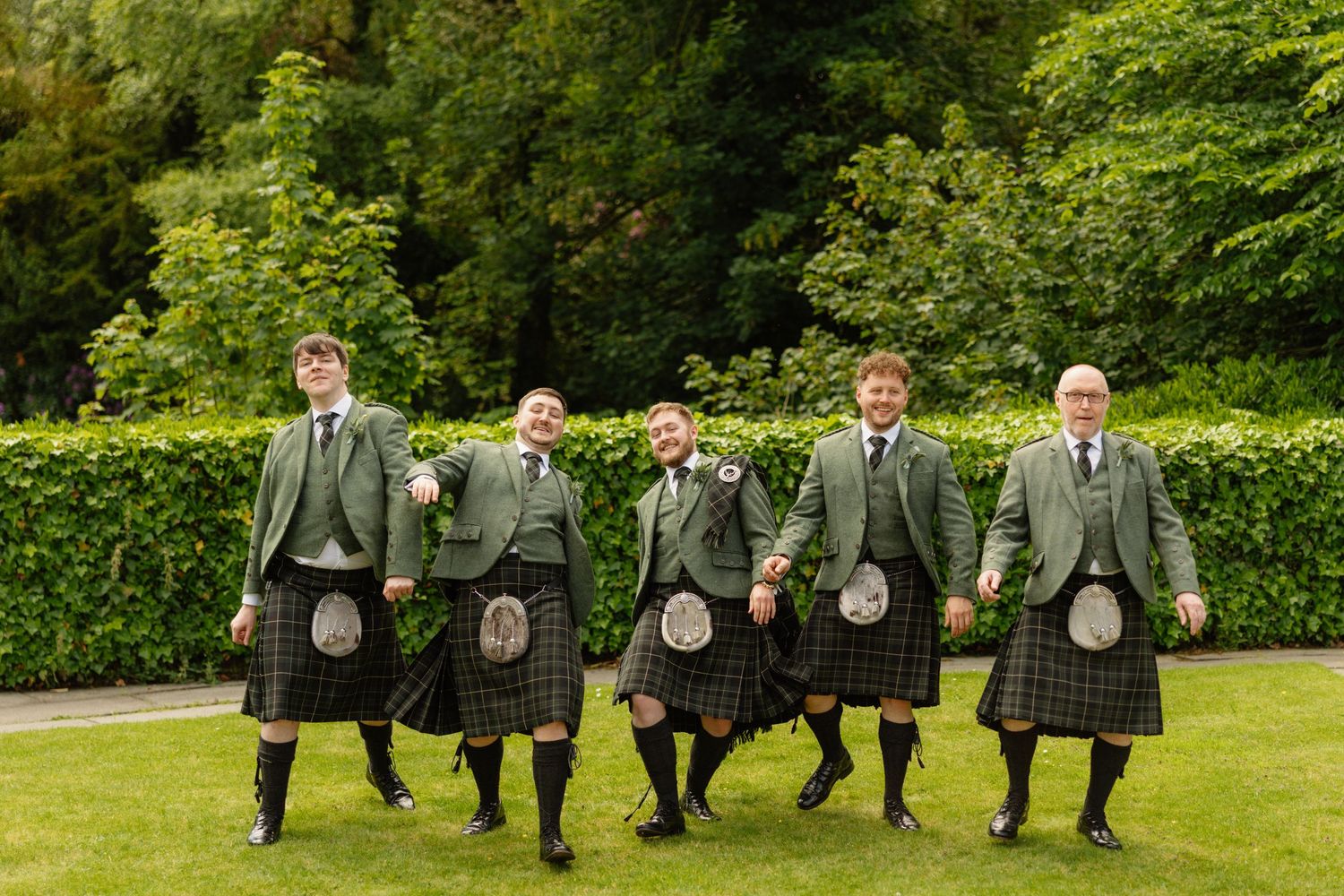 Five men in matching green tartan kilts and jackets walk together outdoors on a lawn at a Scottish wedding.