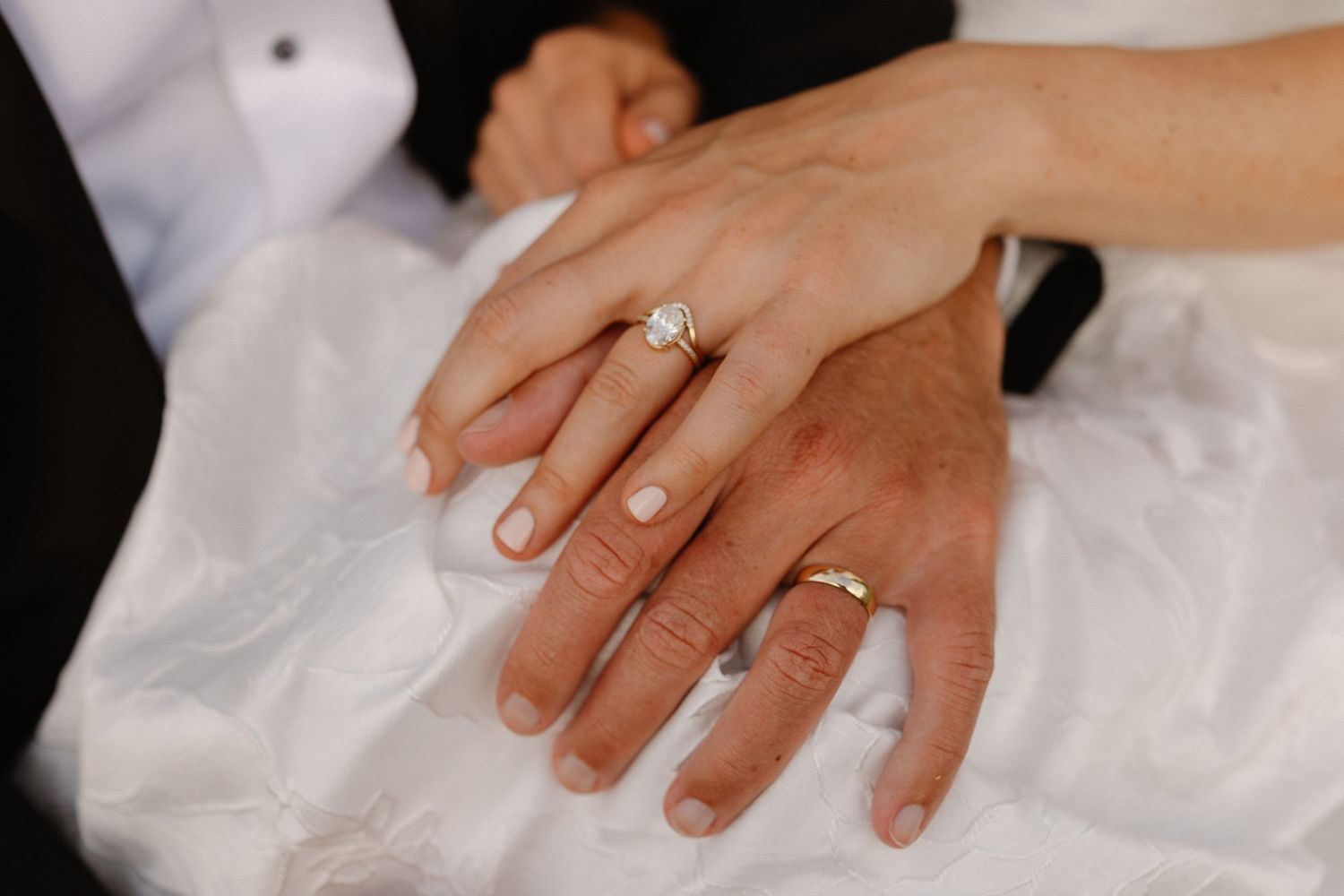 Close-up of bride and groom hands showing diamond engagement ring and gold wedding band on white dress.