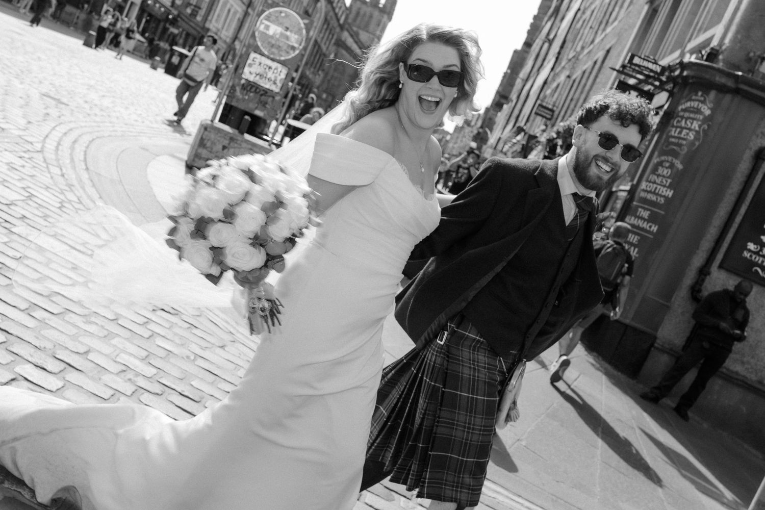 Black and white photo of laughing bride and groom in kilt on cobblestone street.