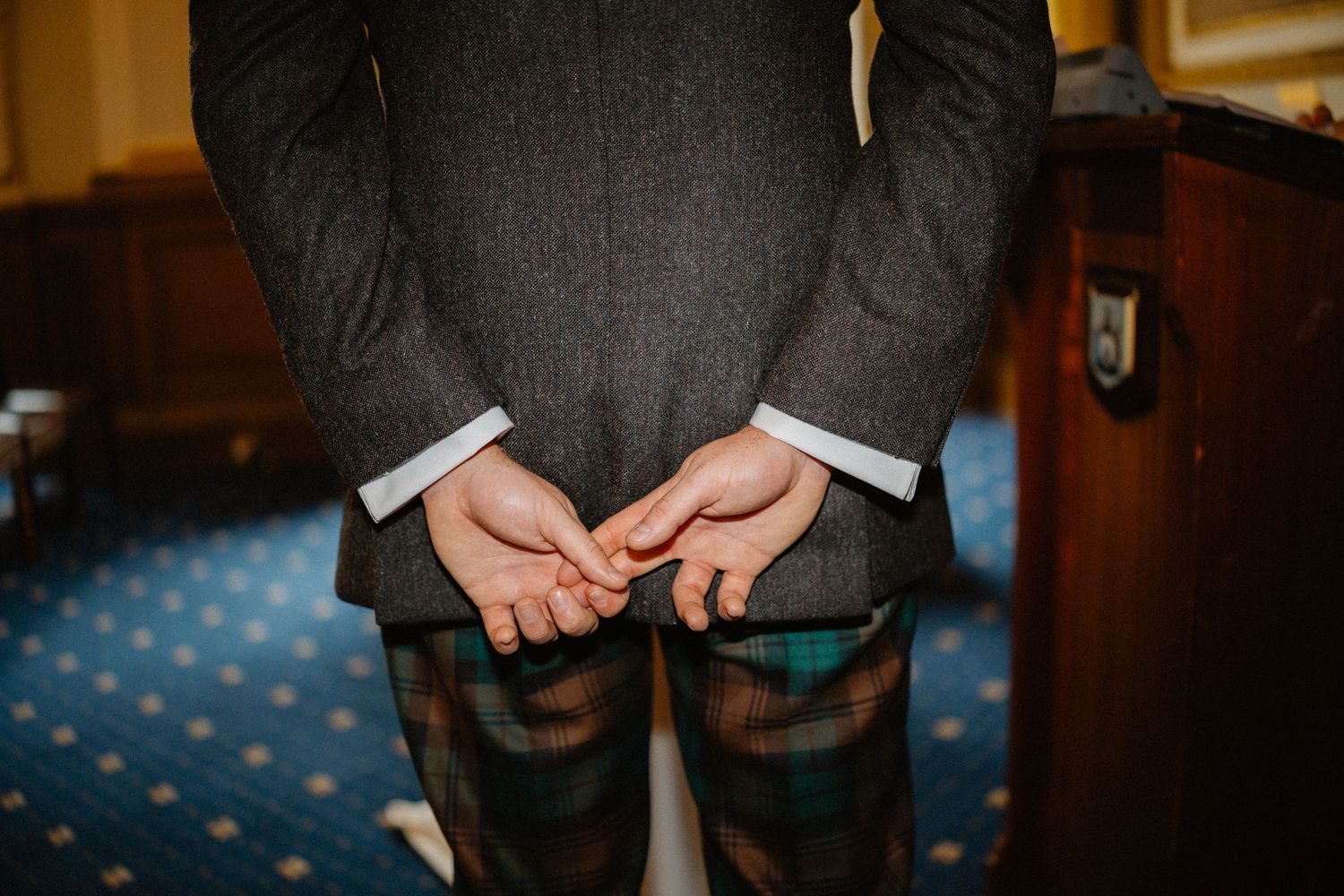 Person in gray suit jacket and tartan kilt standing with hands clasped behind back in elegant room.