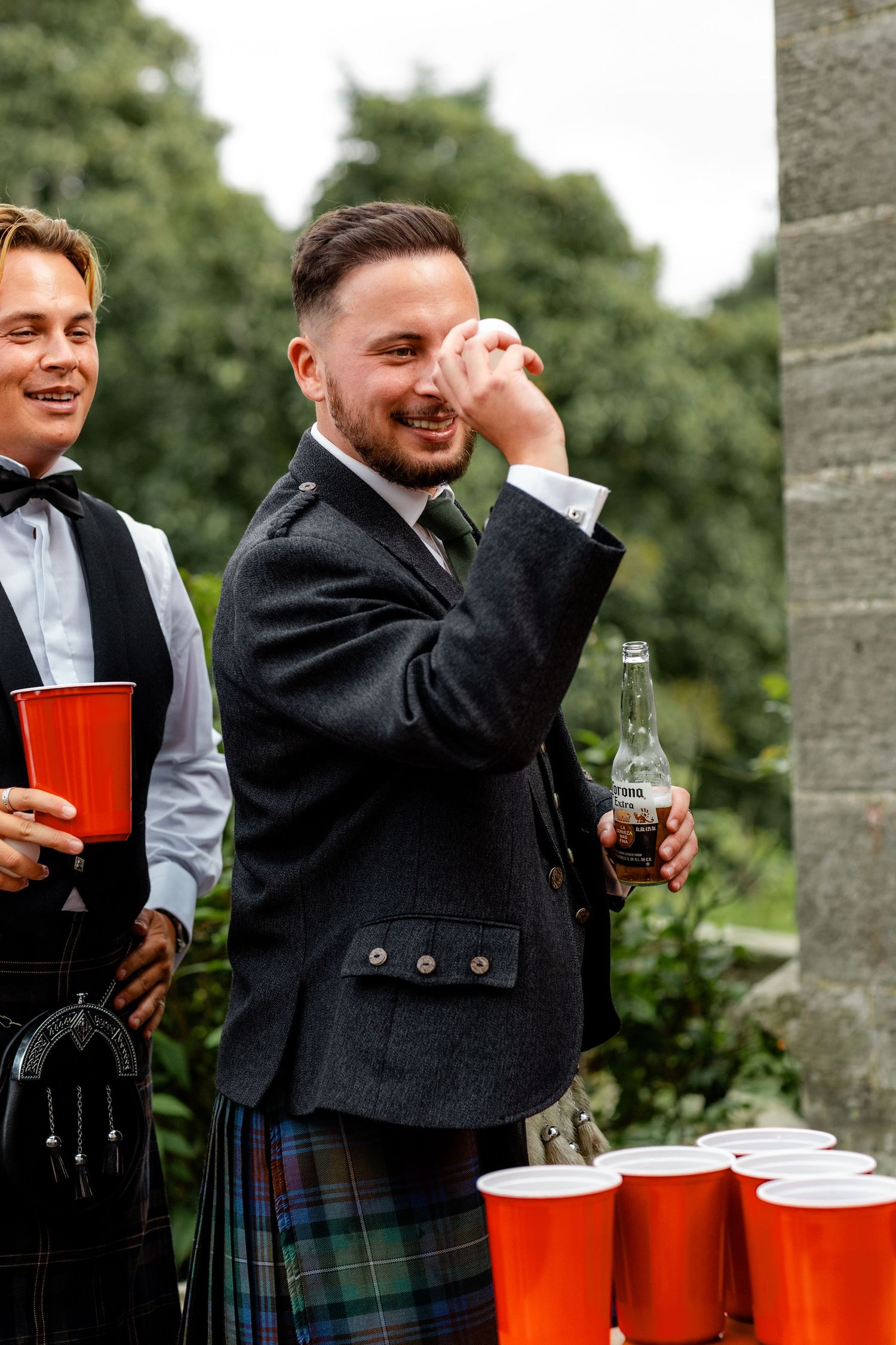 A smiling man in a Scottish kilt and jacket holds a bottle while playing beer pong at a wedding celebration.