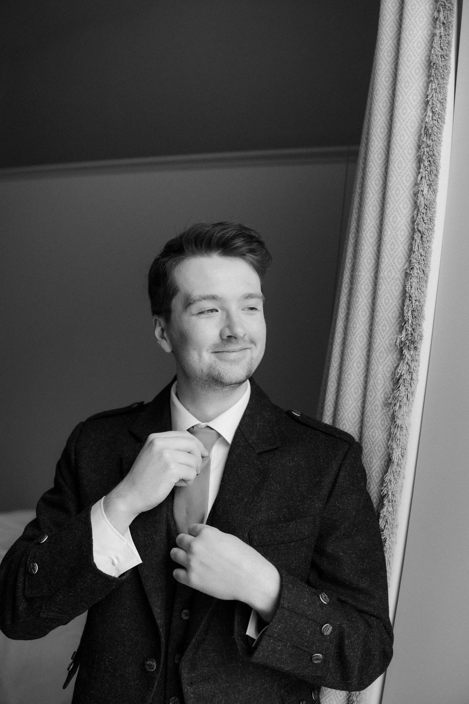Groom in a dark suit adjusting his tie while smiling in a black and white wedding portrait.