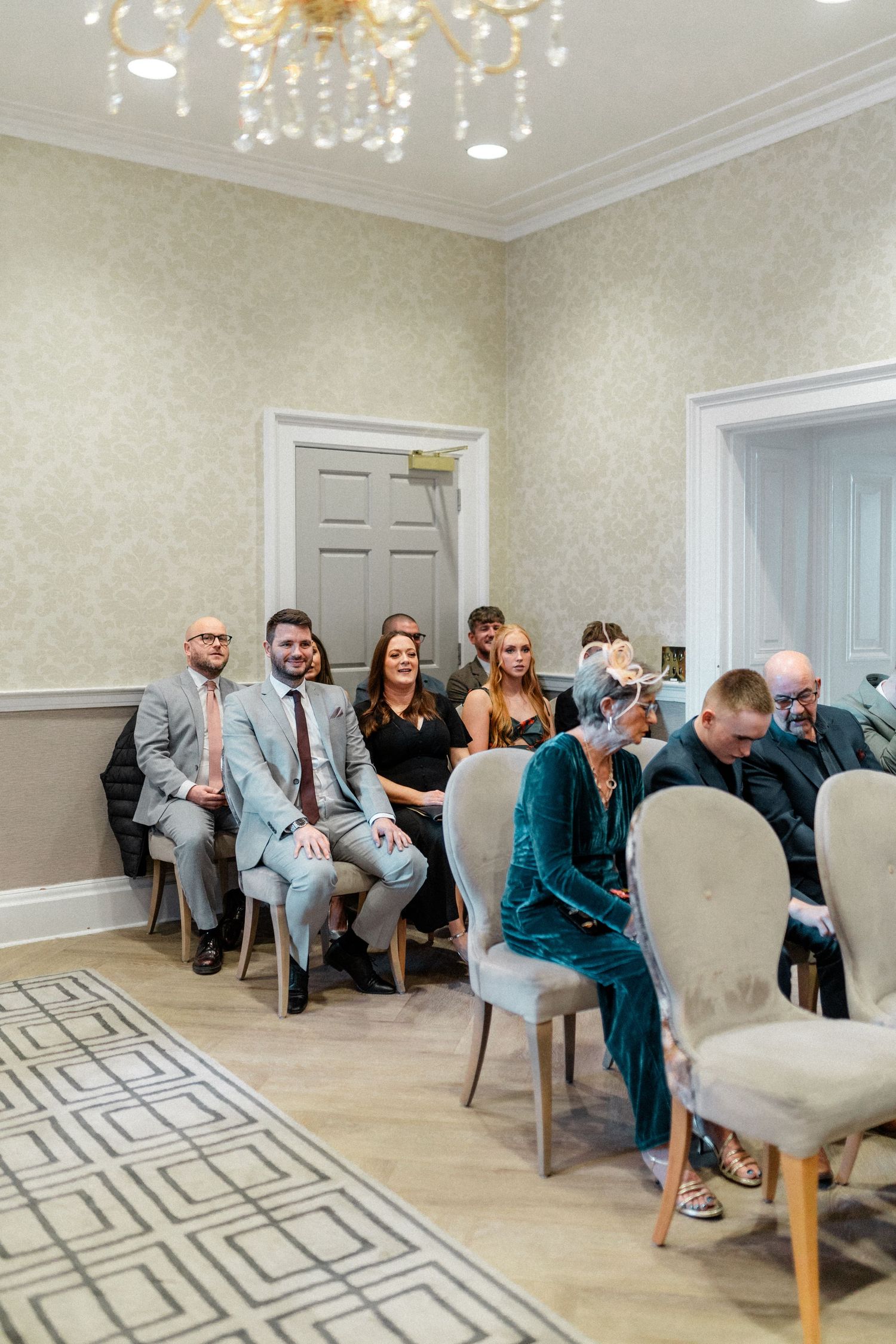 Smiling wedding guests seated in elegant cream chairs in a decorated ceremony room with chandelier.