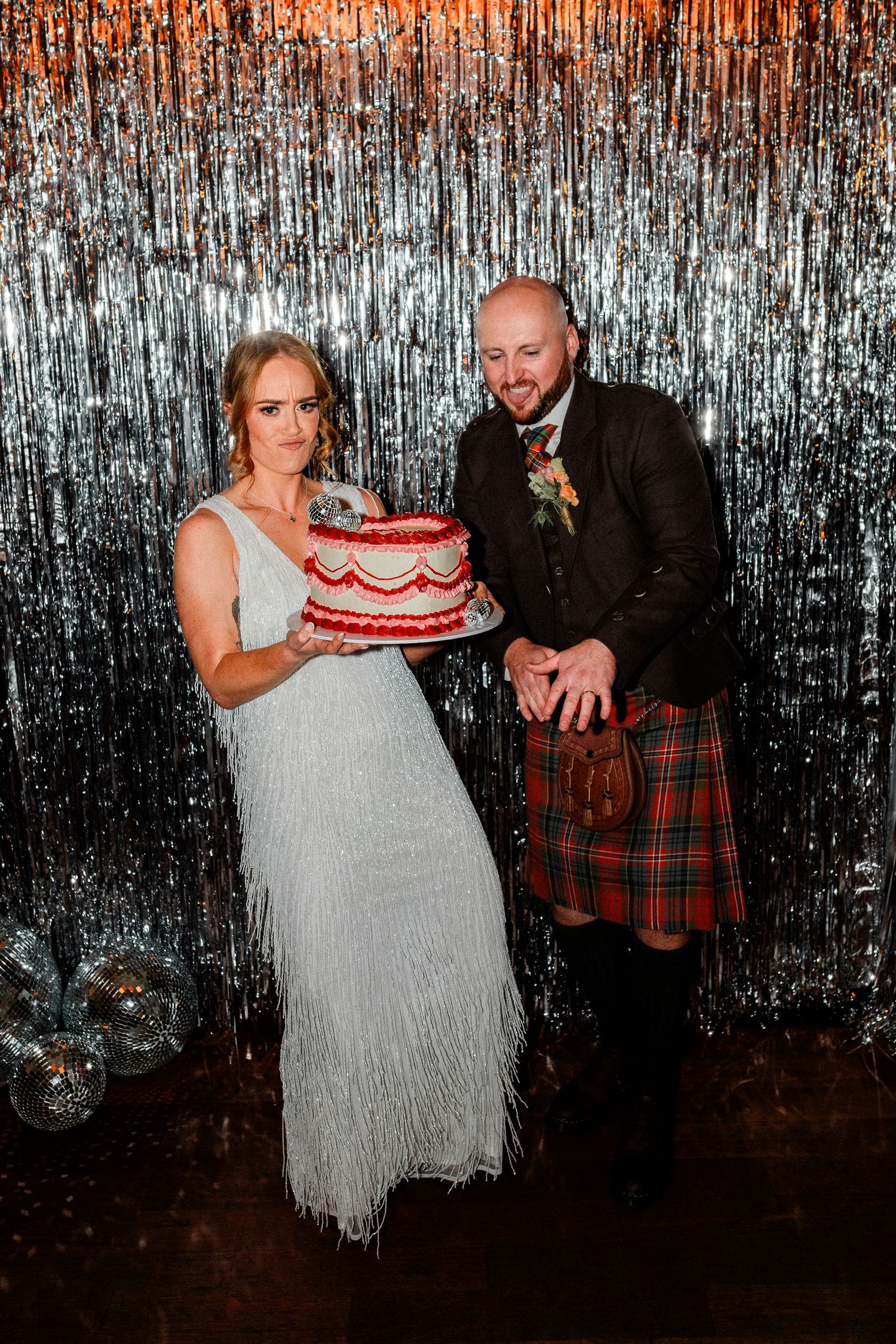 Bride and groom holding red velvet wedding cake in front of silver tinsel backdrop.