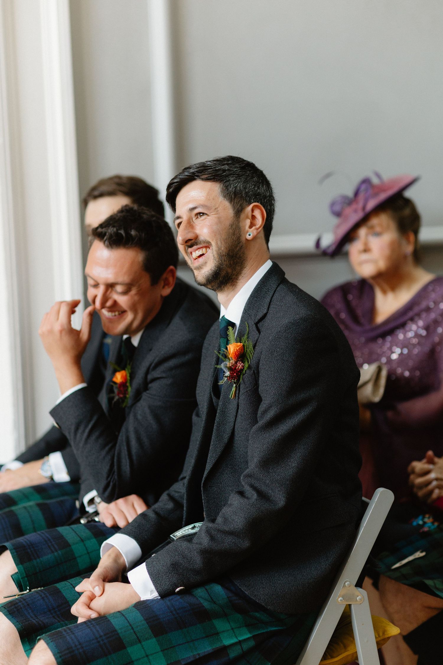 Groom in dark suit with floral boutonniere laughing joyfully during Scottish wedding ceremony with guests in tartan kilts.