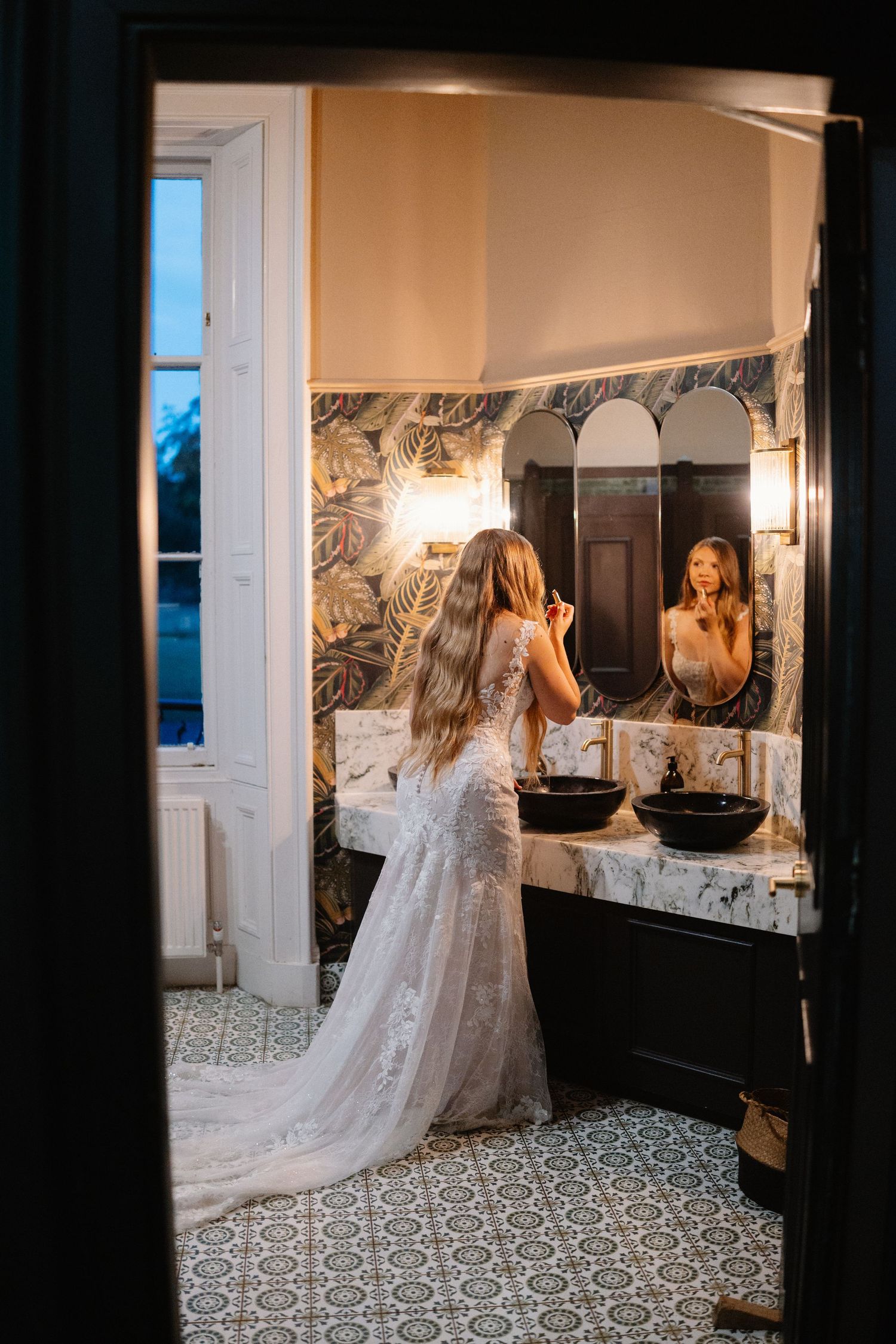 Bride in a flowing lace gown admires her reflection in an elegant bathroom with patterned floor tiles.
