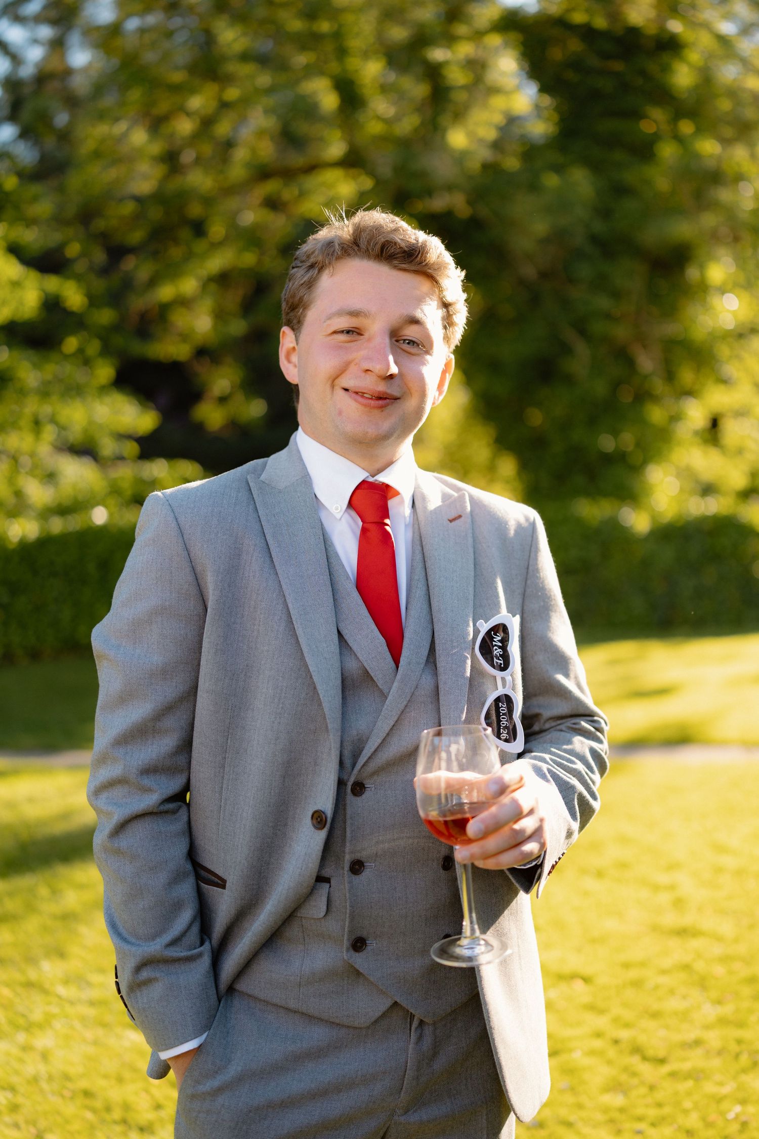 Smiling groomsman in a grey three-piece suit with red tie holding a rose wine glass outdoors in sunlight.
