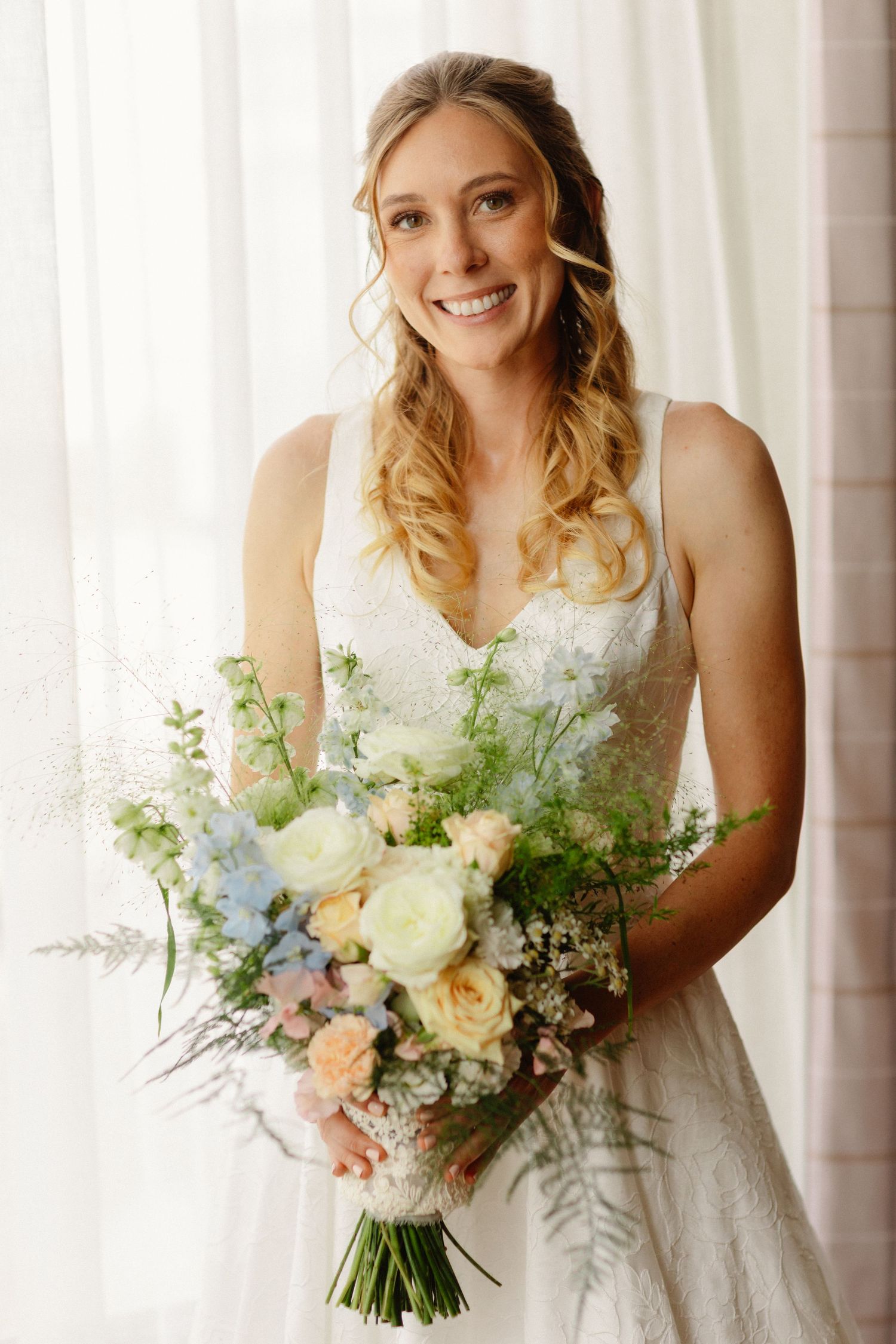 Smiling bride in white lace gown holds pastel bouquet of roses and wildflowers, standing near sheer curtains.