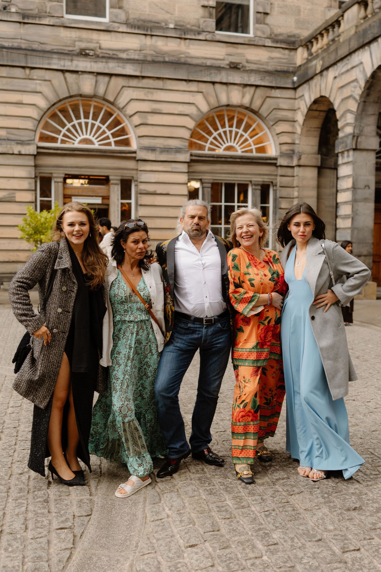 Group of five elegantly dressed guests posing together in a cobblestone courtyard outside a historic stone building.