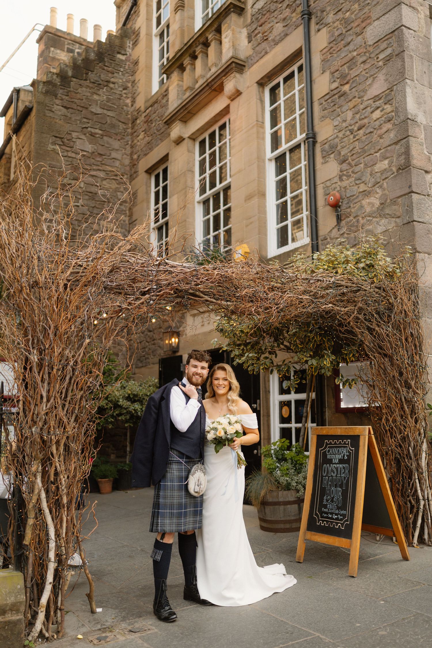 A bride and groom pose outside a historic stone building, groom wearing a traditional Scottish kilt and sporran.