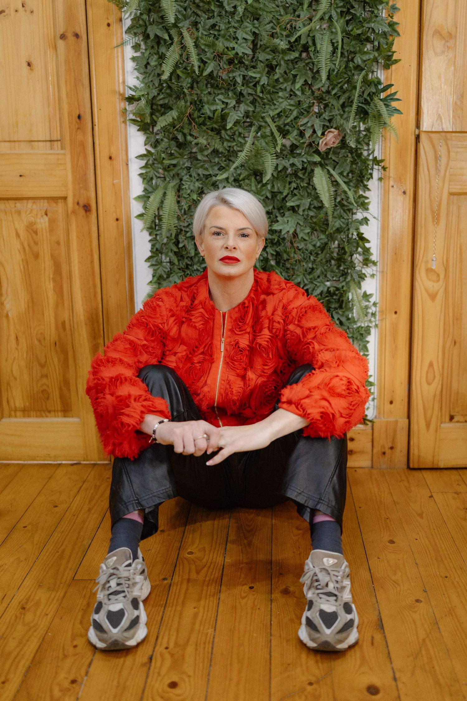 A person in a bright red lace top and dark pants sits on wooden floor against a green foliage backdrop.