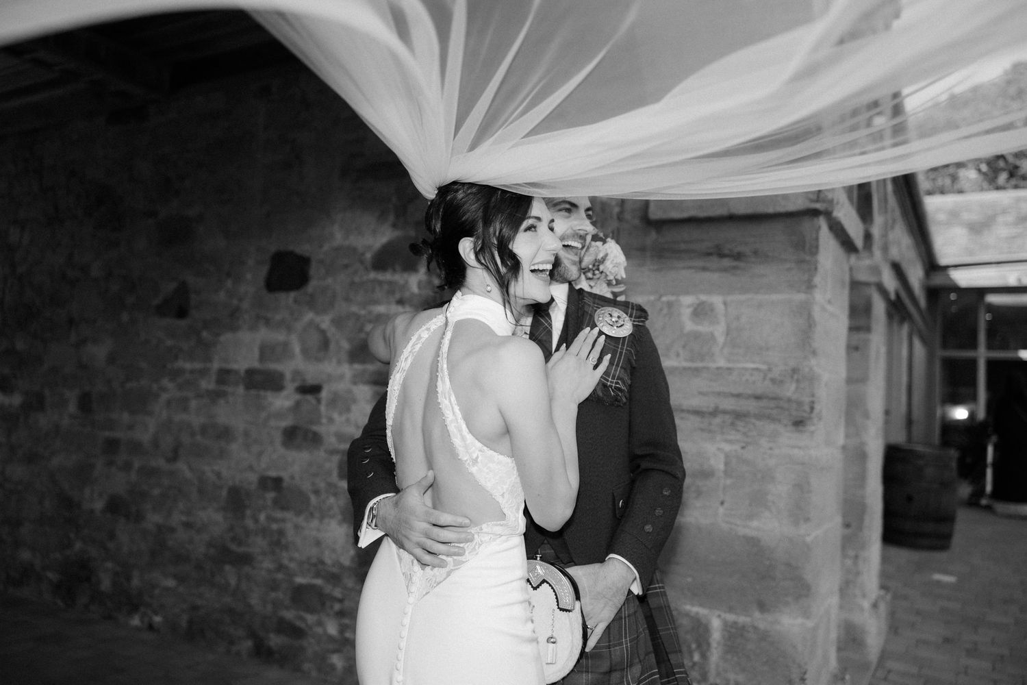 Wedding couple embraces under flowing white veil creating romantic black and white portrait.