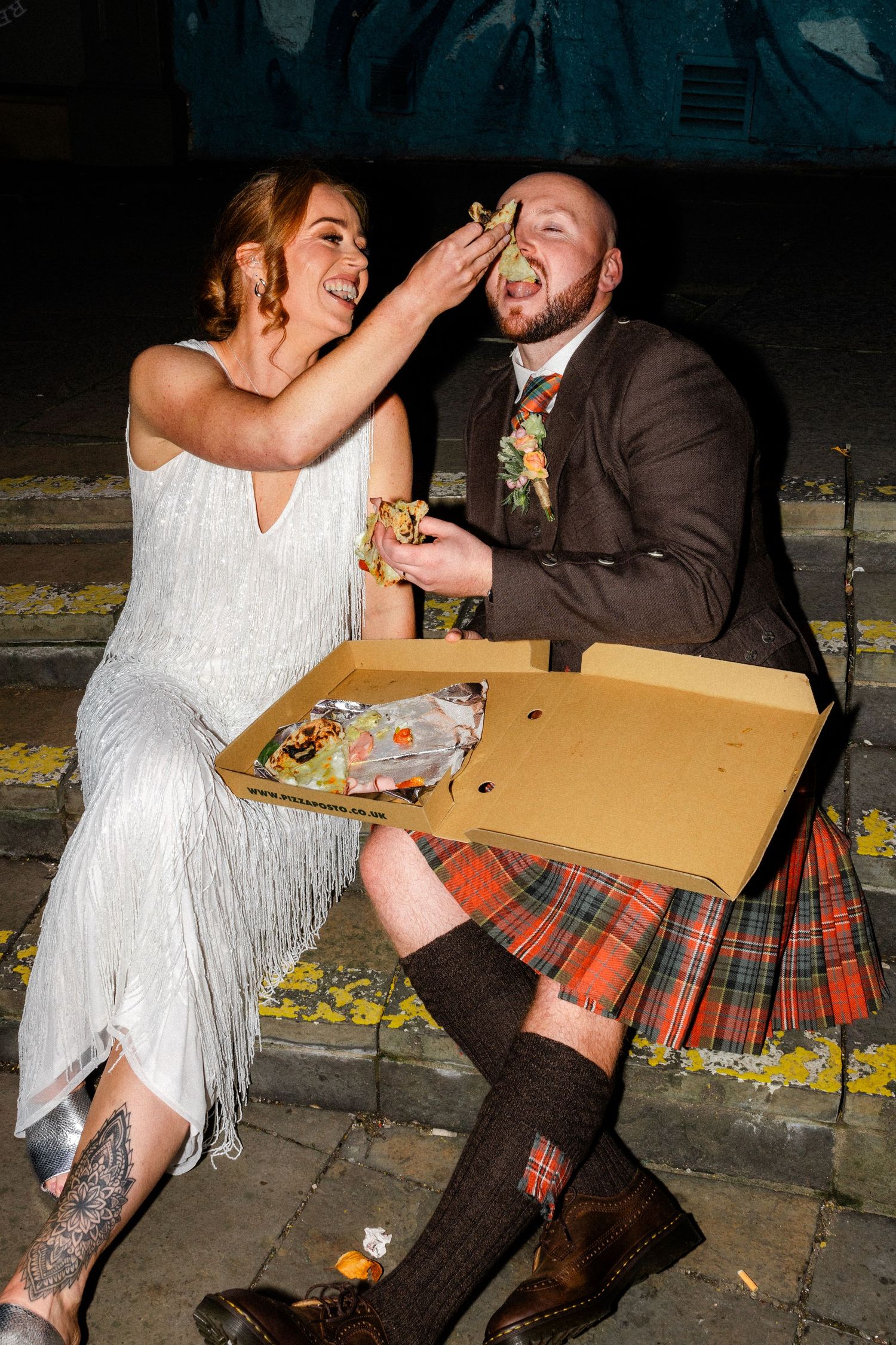 A couple in wedding attire sharing pizza on a street curb at night.