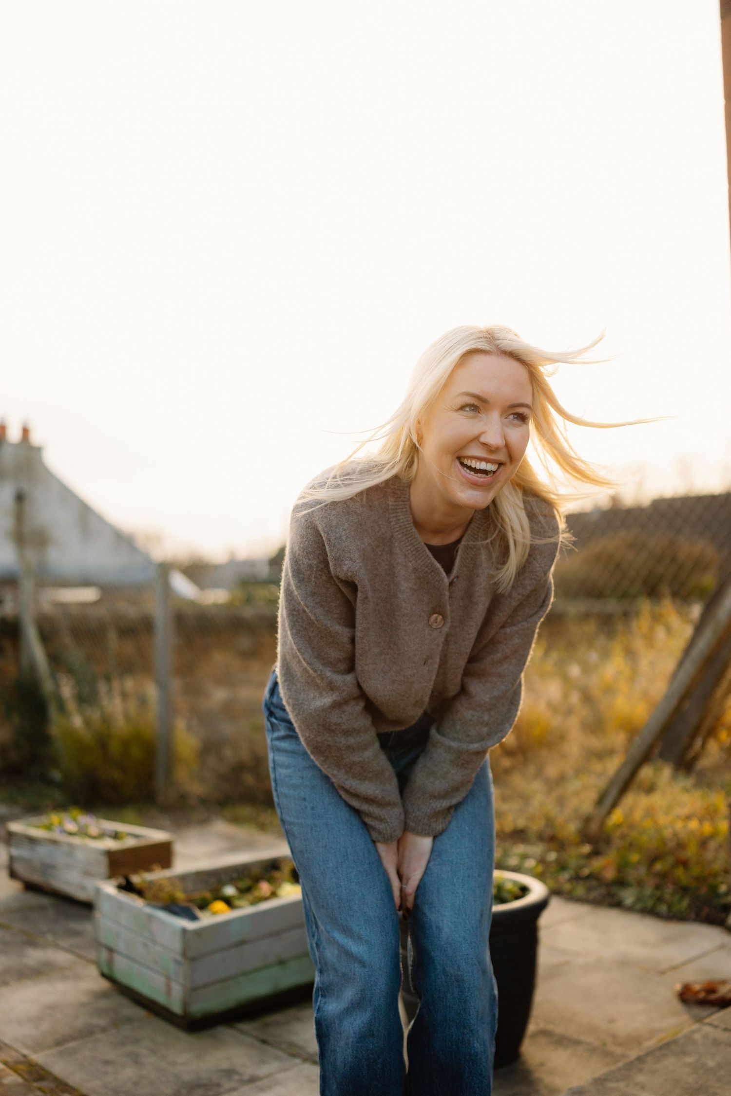 Person in brown sweater and jeans laughing joyfully in an outdoor garden setting at sunset.