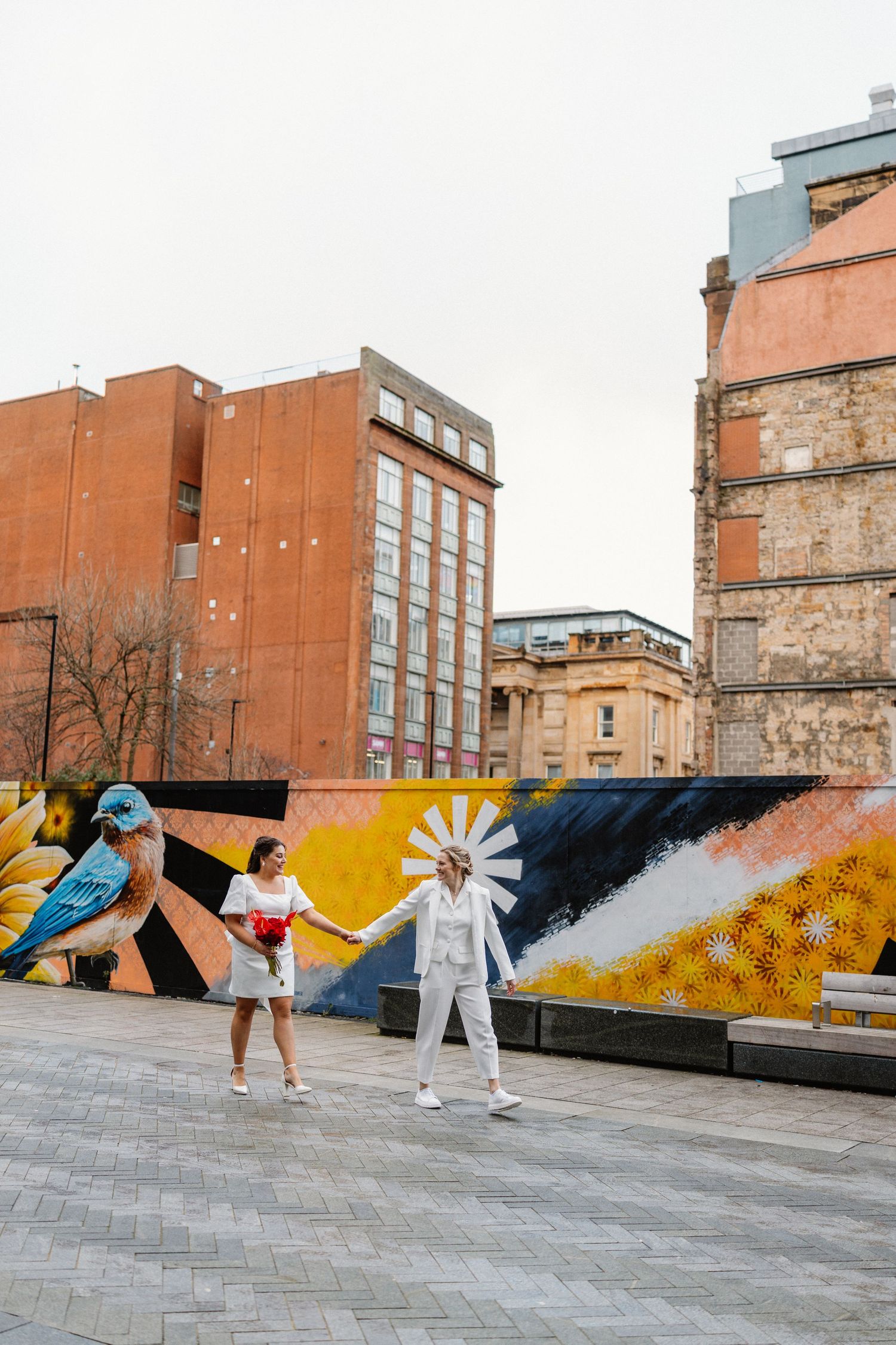 Two brides in white hold hands before a colorful street mural in an urban setting.