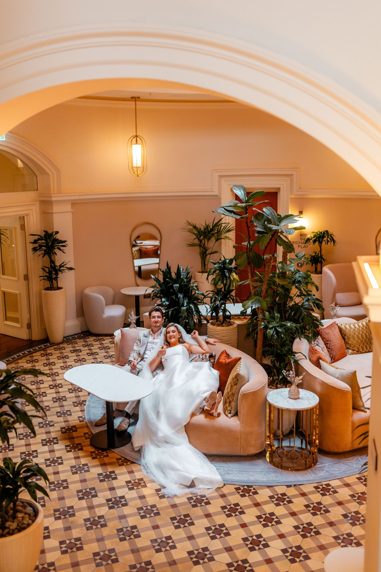 Bride and groom relaxing on a elegant sofa in a warmly lit hotel lobby with arched ceilings and lush plants.