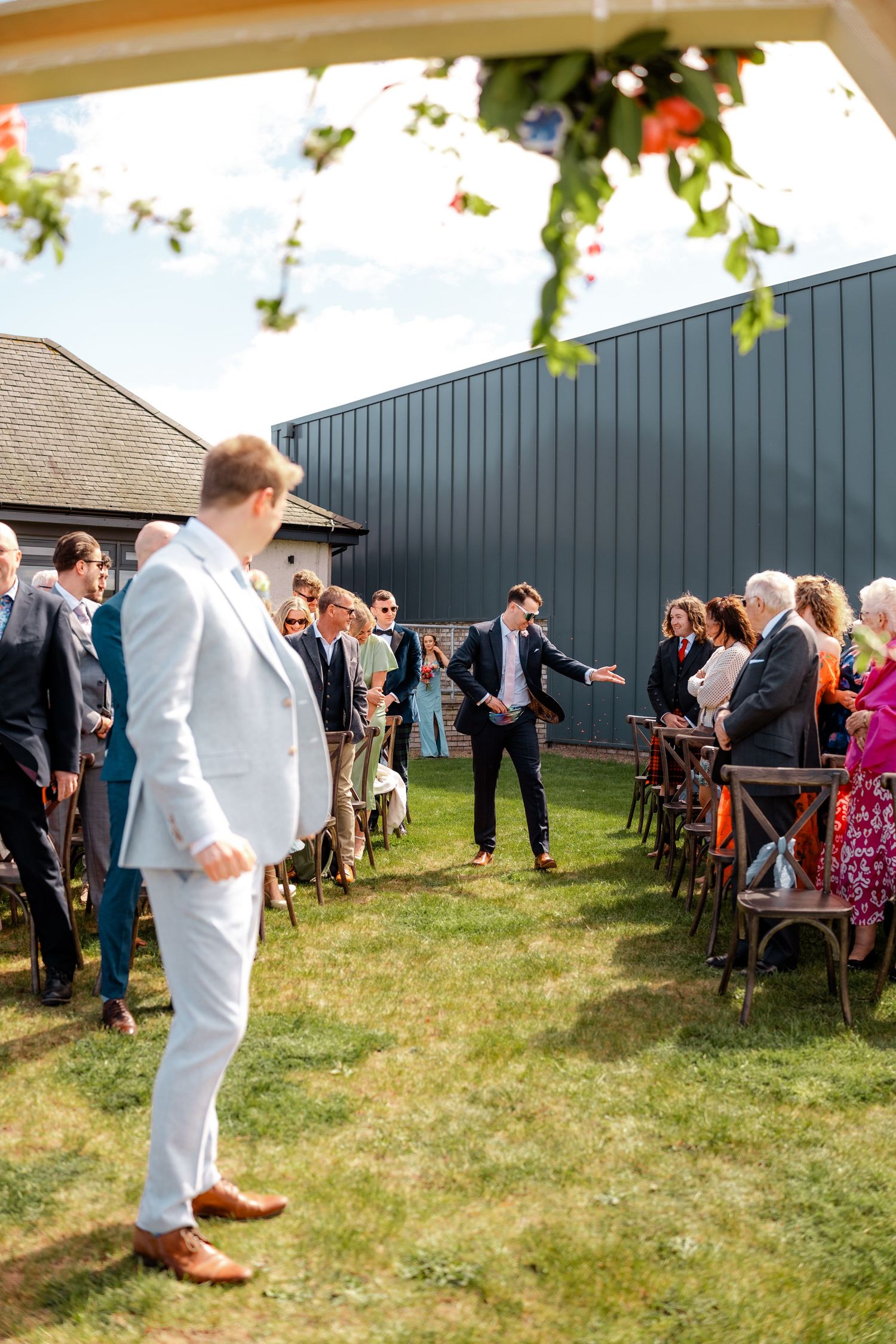 Groom in light blue suit waits at outdoor wedding ceremony aisle beside dark barn venue with seated guests.