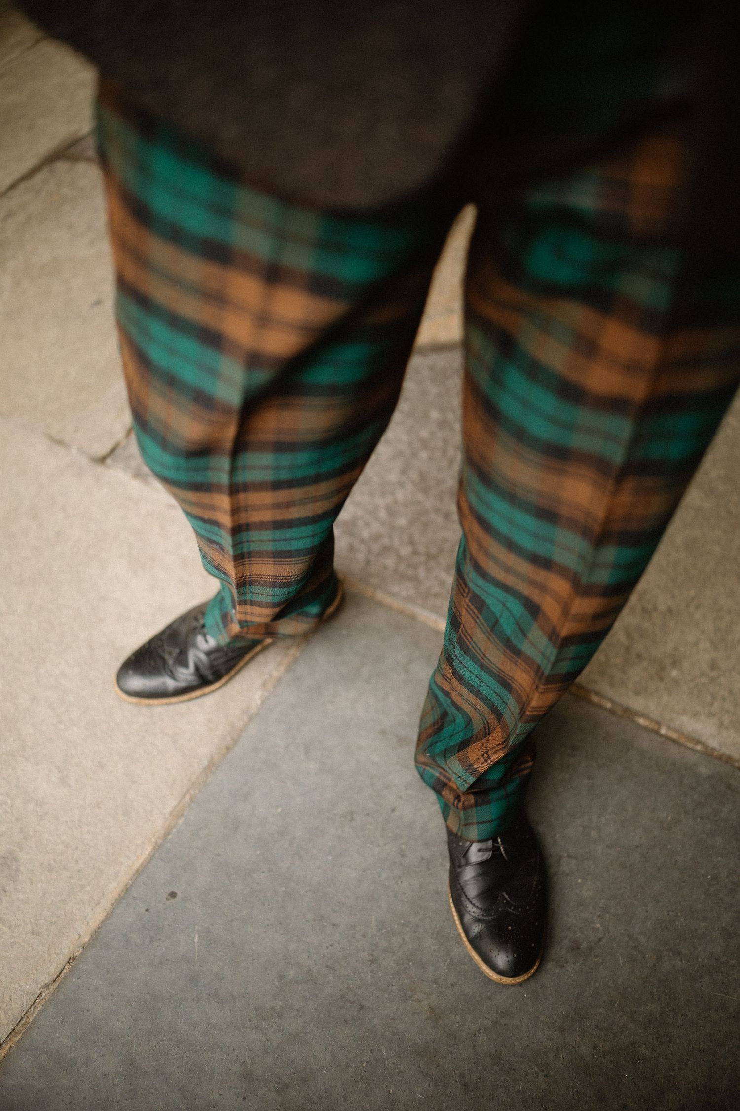Close-up of man wearing green and brown tartan plaid trousers with black leather dress shoes on stone pavement.