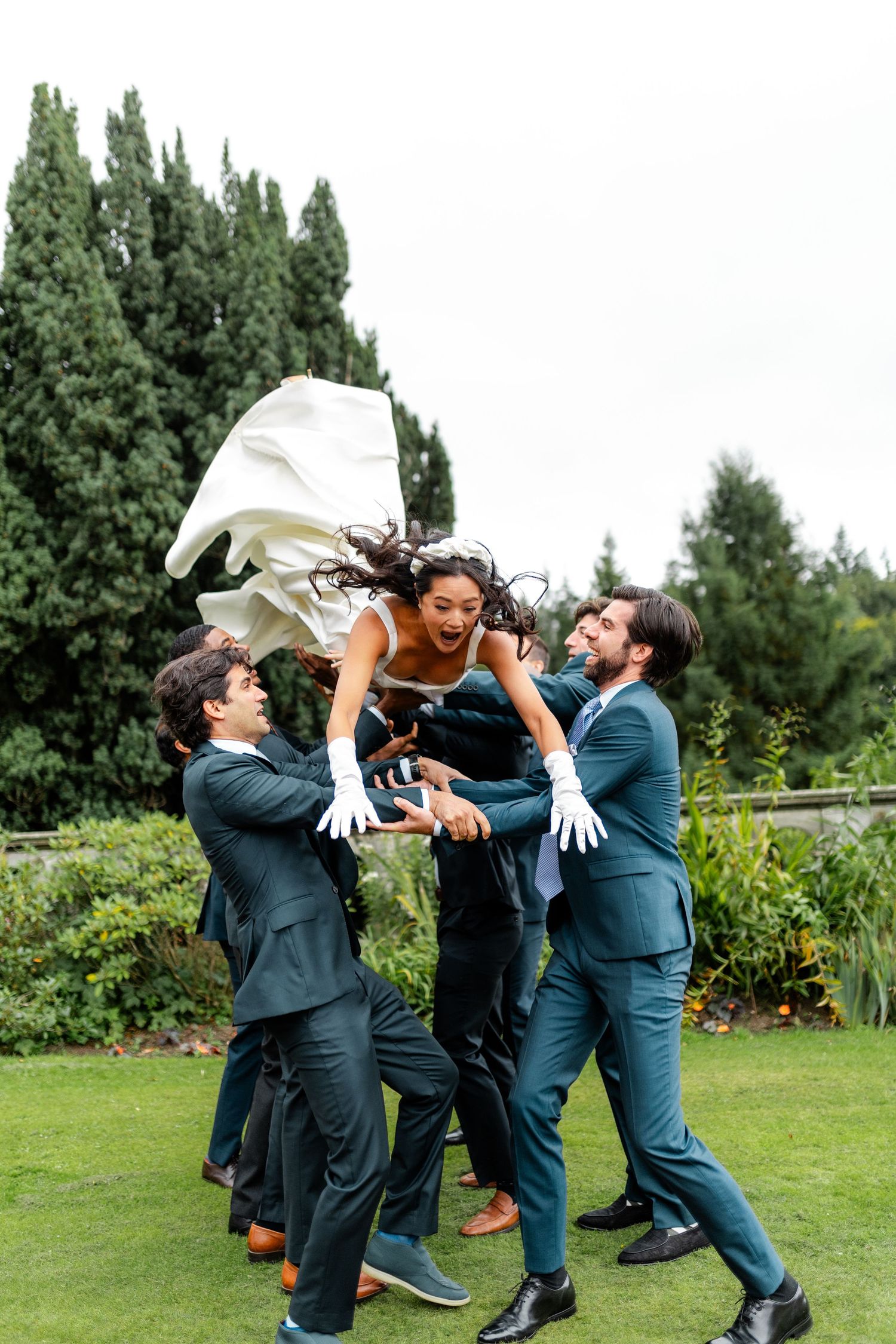 Groomsmen in teal suits toss a laughing bride into the air outdoors at a wedding celebration.
