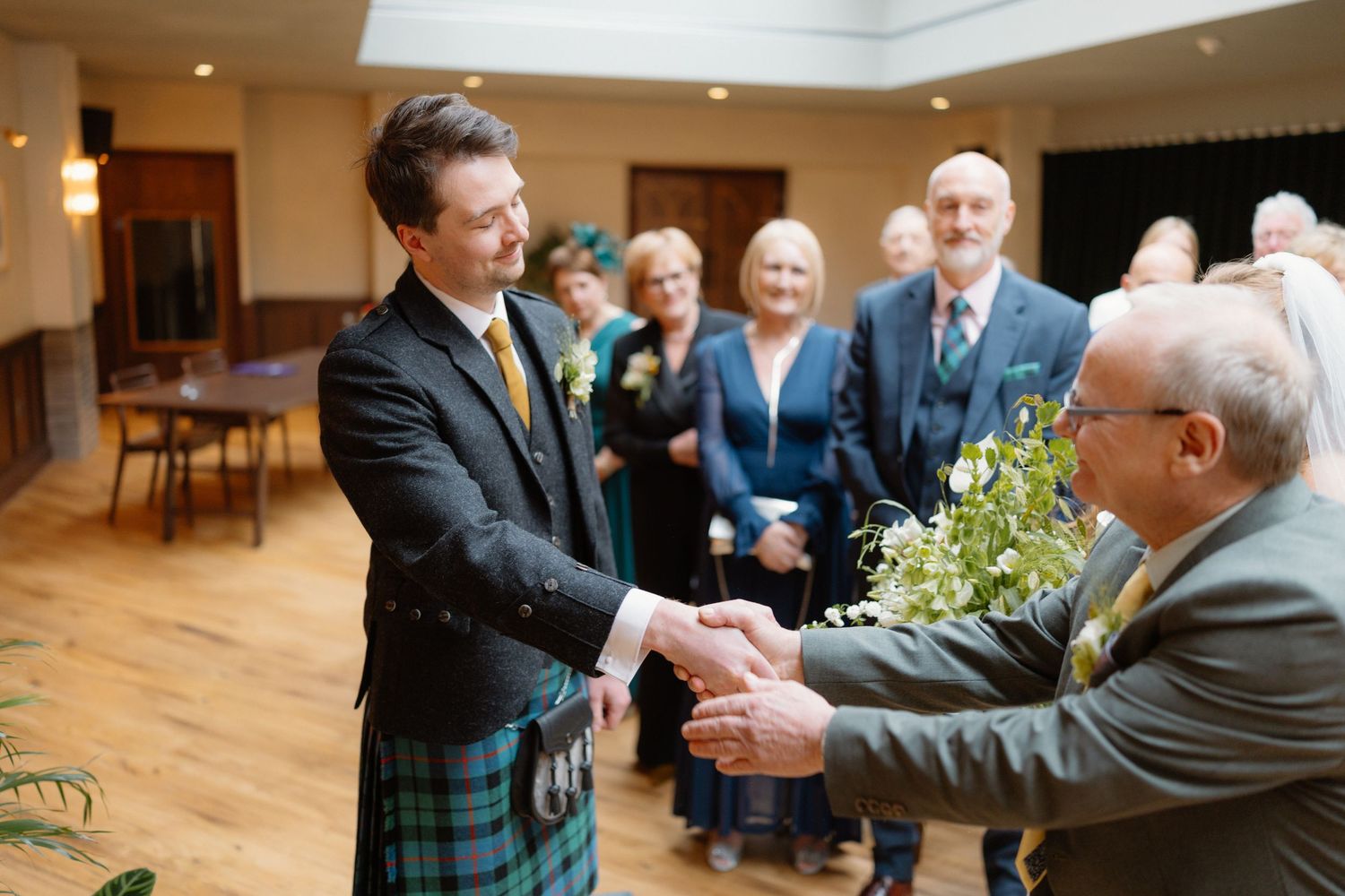 Groom in Scottish tartan kilt shaking hands with wedding guest during reception ceremony.