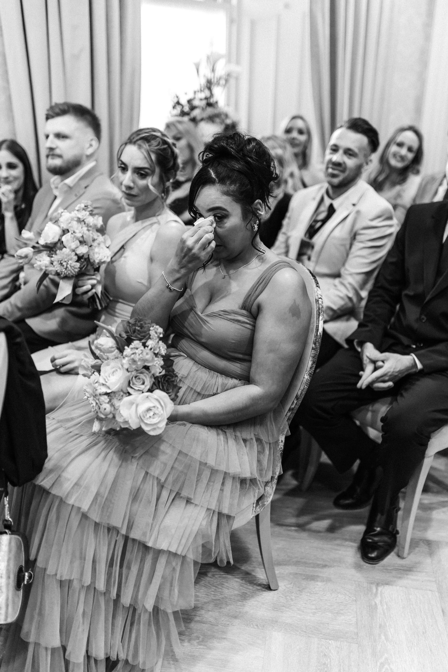 Emotional bridesmaid wipes tears during wedding ceremony in black and white photo.