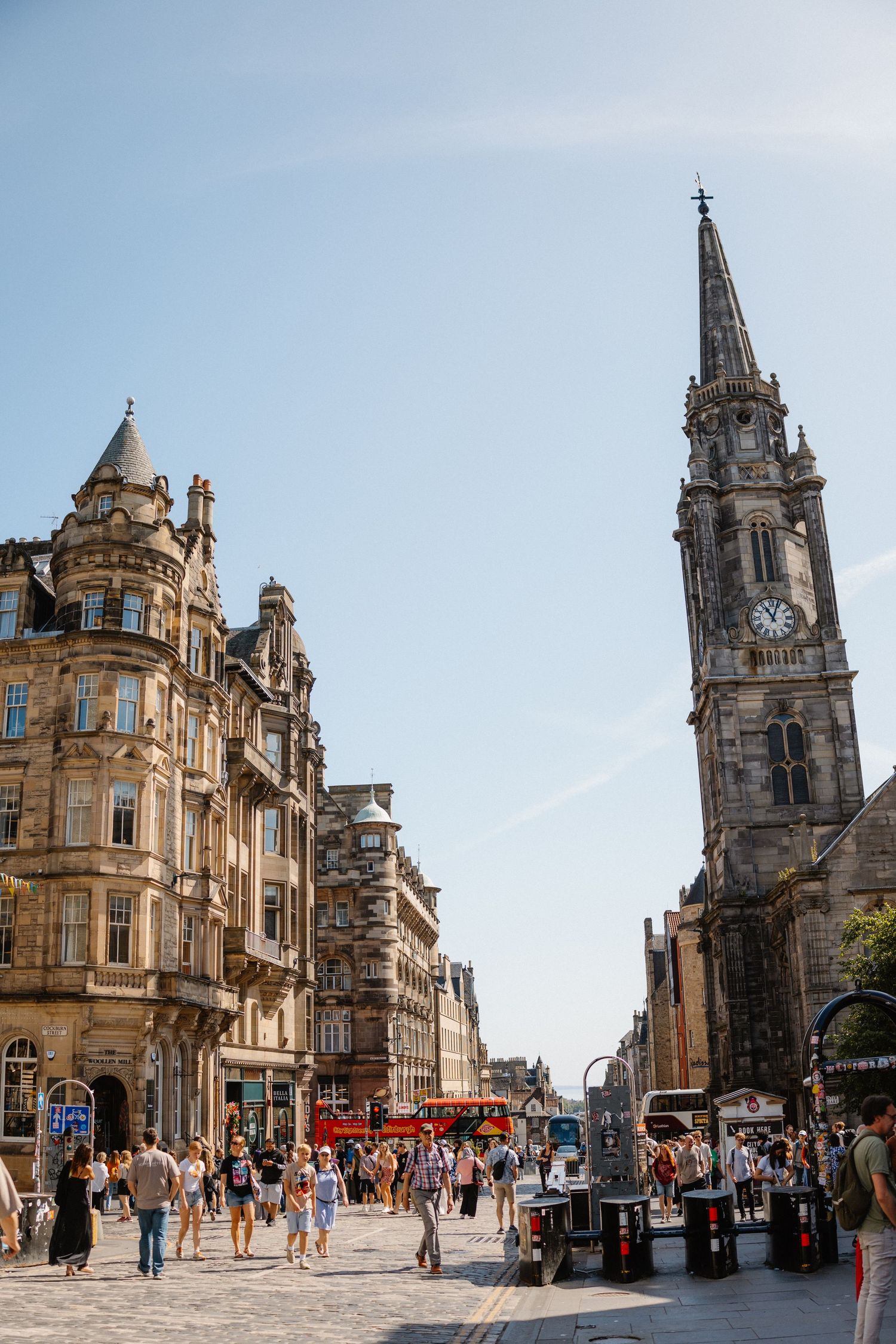 Busy Royal Mile street in Edinburgh with historic stone buildings, a church steeple, and tourists on a sunny day.