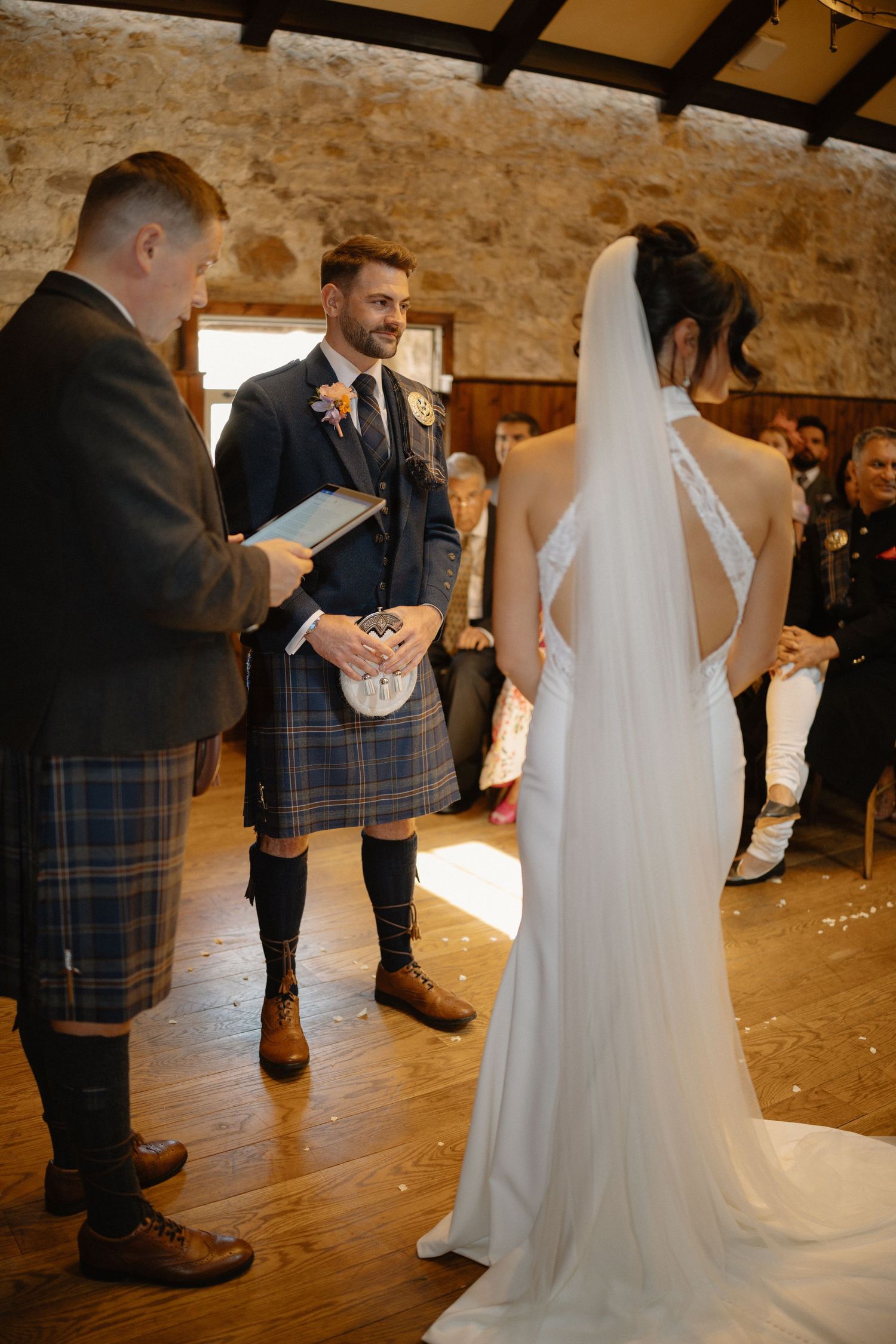 Groom in traditional Scottish tartan kilt smiles at bride during indoor wedding ceremony in stone venue.