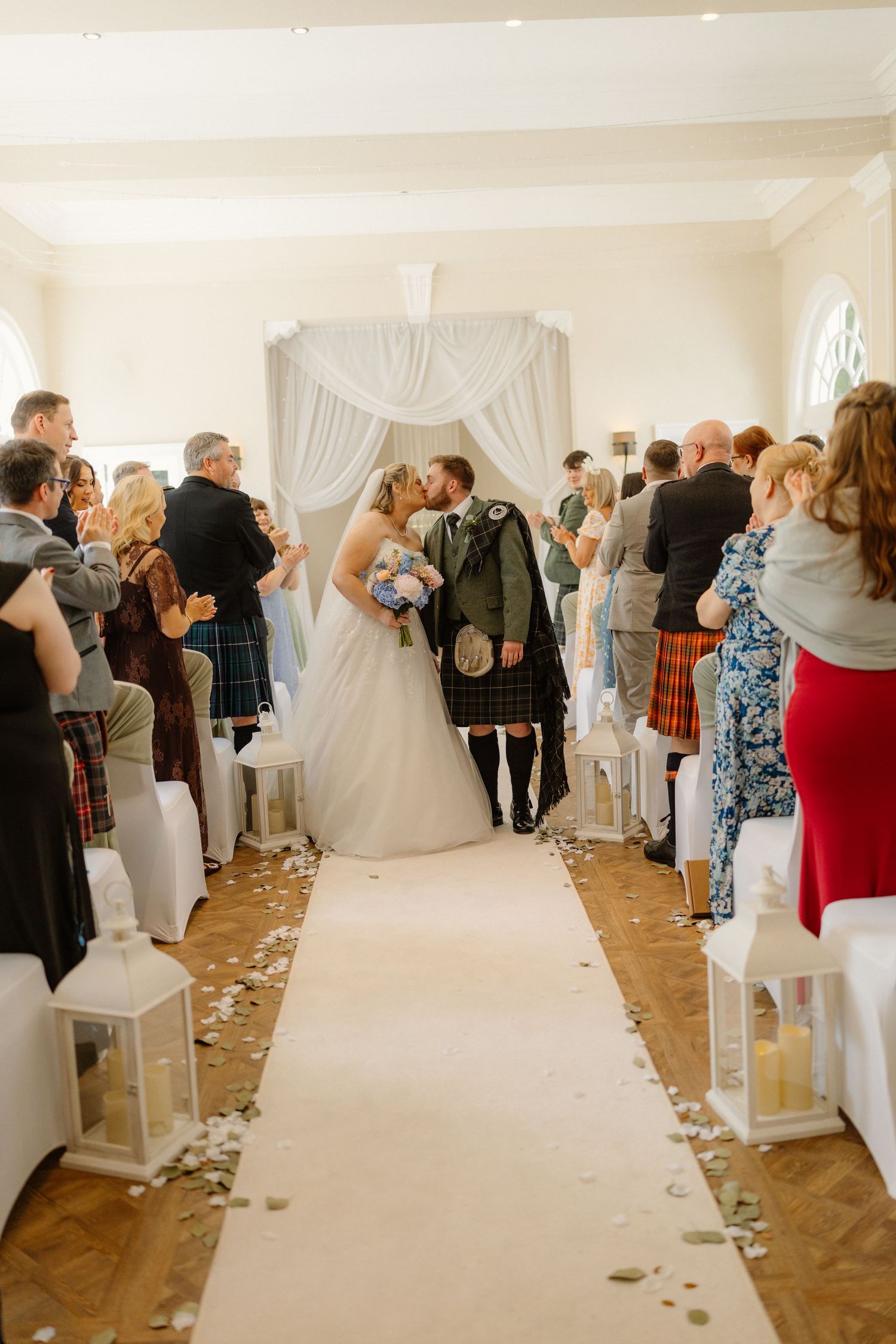 Bride and groom share first kiss at altar while wedding guests applaud in elegant ceremony hall.