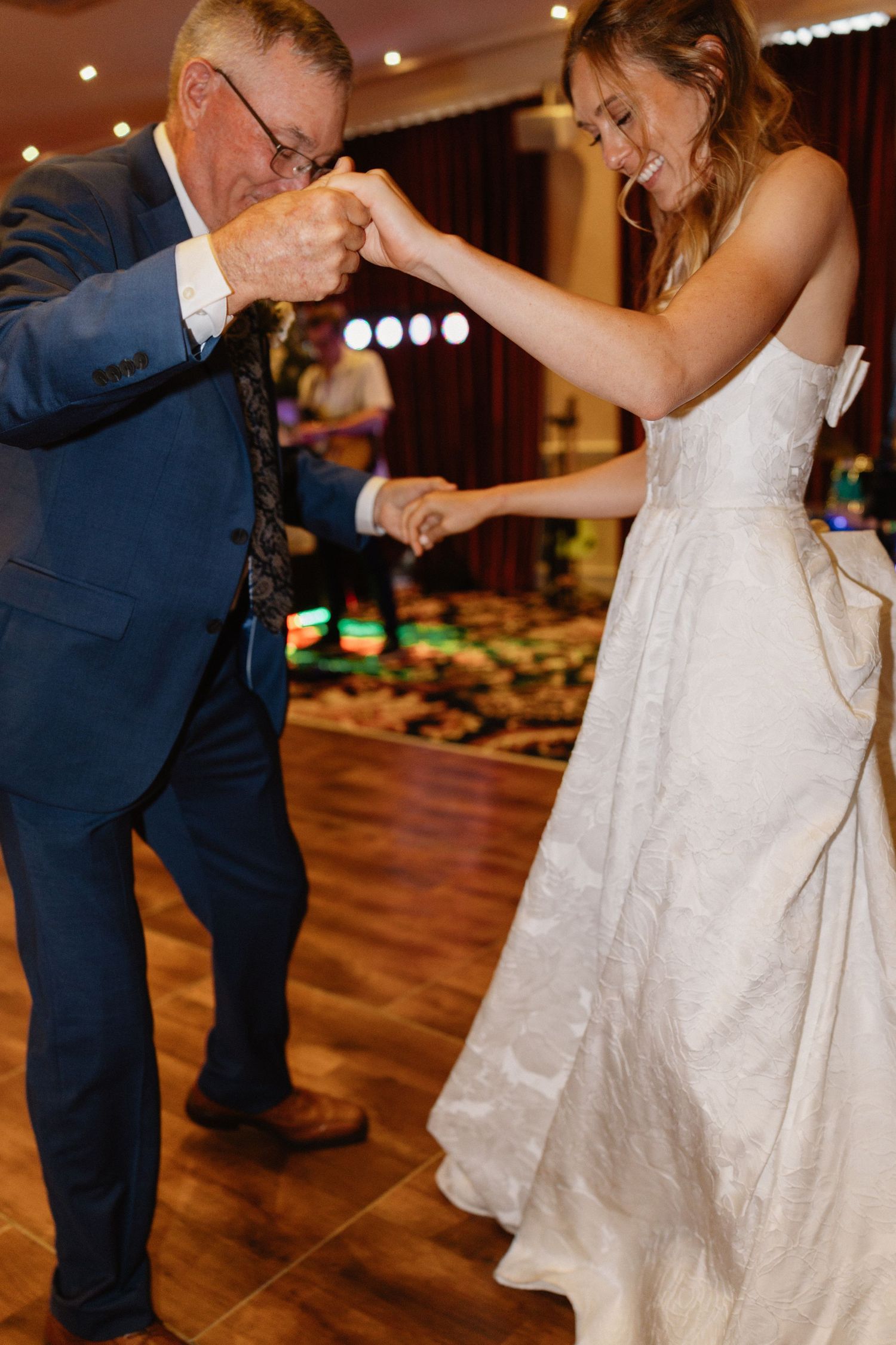 Father kisses bride's hand during emotional father-daughter dance at wedding reception.