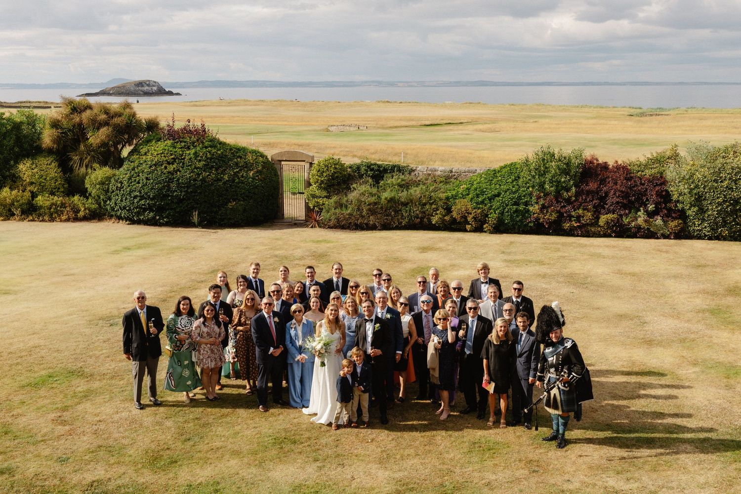 Large wedding group photo outdoors on a sunny day with ocean and island views in the background.