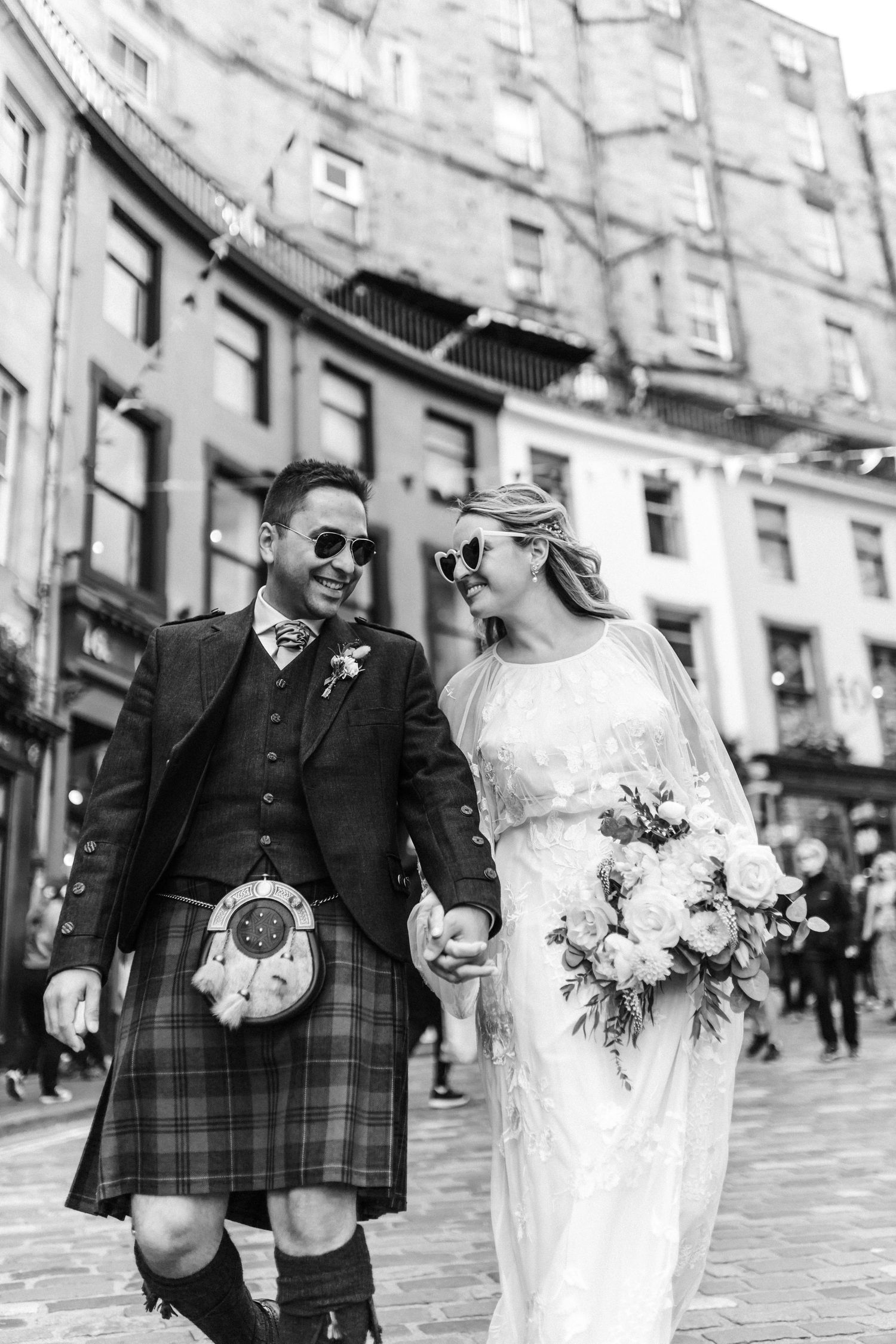 Black and white photo of a smiling bride and groom in Scottish kilt walking hand in hand on cobblestone street.