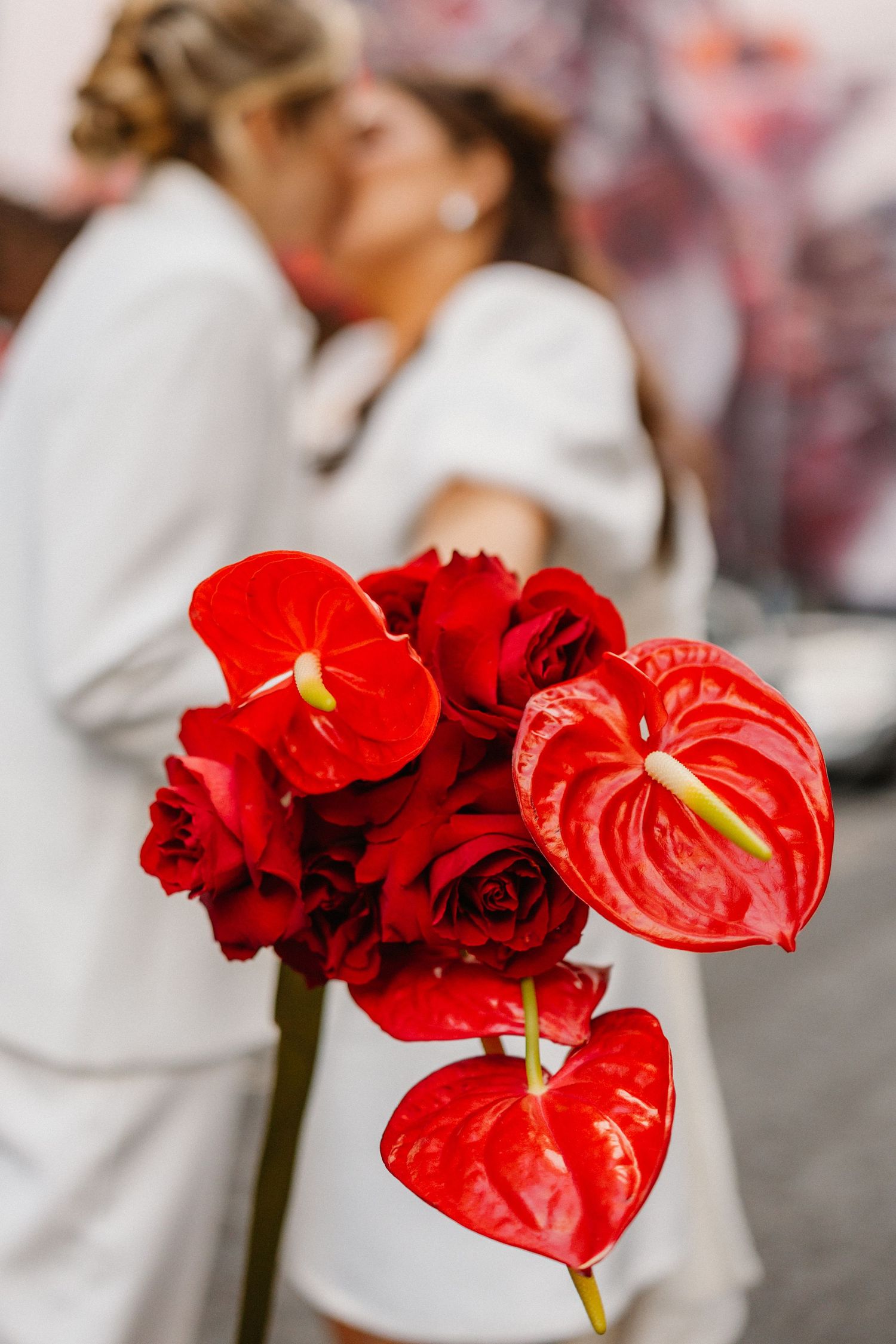 Vibrant red anthurium and rose wedding bouquet held in focus, with a couple kissing in white outfits blurred behind.