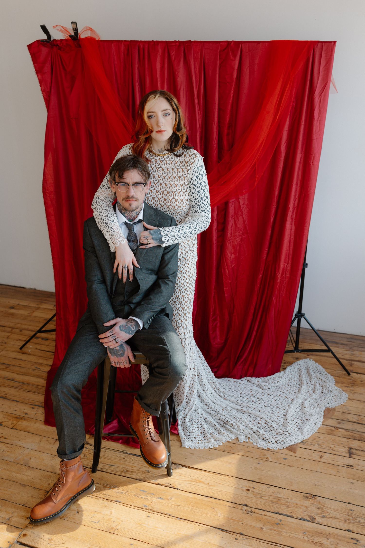Couple poses before red drape backdrop, bride in lace gown stands behind groom in dark suit seated on chair.