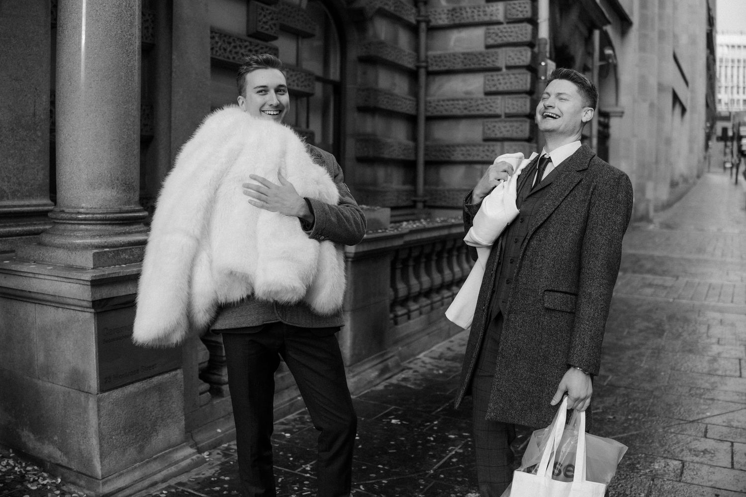 Black and white photo of two men laughing on a city street, one wearing a fur coat.